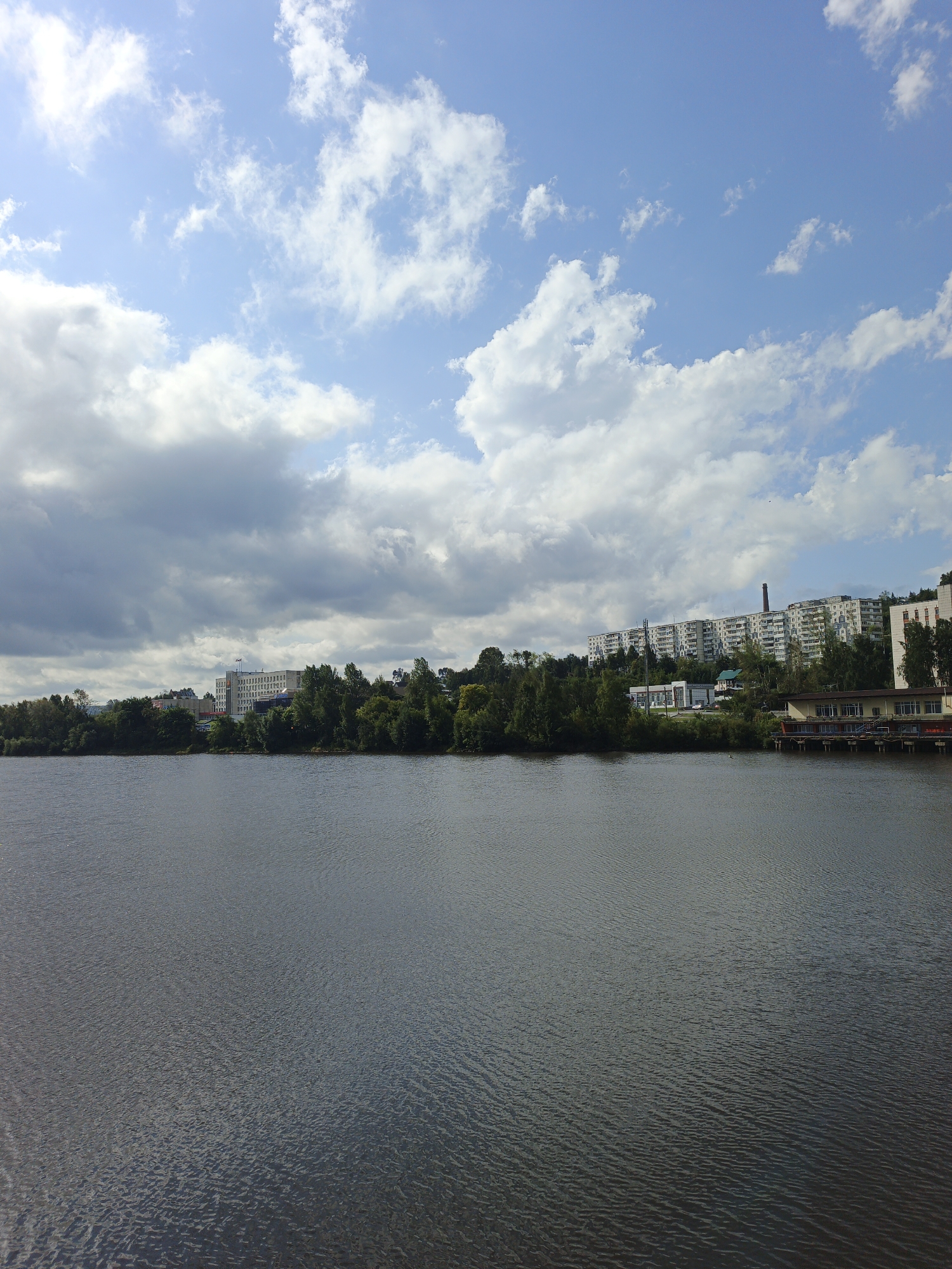 The image depicts a wide, calm body of water, likely a river or lake, in Zlatoust, Russia. The water's surface is dark and slightly rippled, reflecting the overcast sky. Across the water, a densely treed shoreline is visible, with various buildings and structures interspersed. The buildings include several multi-story apartment complexes, some appearing to be Soviet-era architecture with long, uniform facades. A prominent brick smokestack rises above one of the buildings. Other structures include what appears to be a waterfront cafe or restaurant with a pier and tables visible. The sky is a mix of blue patches and white, puffy clouds, with some darker, heavier clouds suggesting a change in weather. The overall impression is a semi-urban landscape bordering a natural body of water, typical of many Russian cities. There are no people visible in the image.
