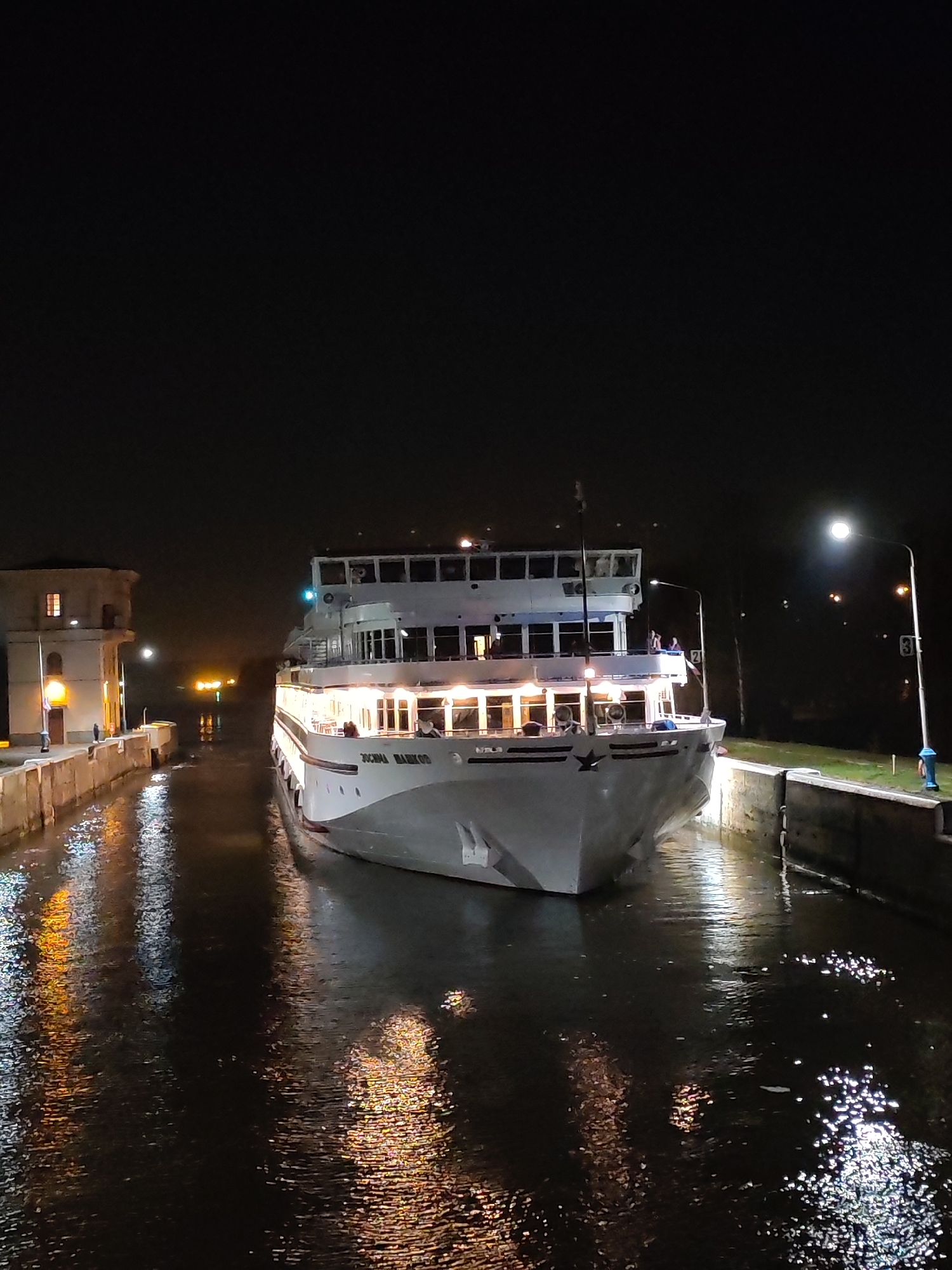 A nighttime shot focuses on a riverboat, named "Zosima Shashkov," navigating a lock in Iksha, Russia. The riverboat is white with multiple decks lit up by interior lights, which reflect in the dark water. The boat is tightly fitted between the concrete walls of the lock. Light posts illuminate the area, casting reflections on the water's surface. There's an adjacent building with a lit window on the left side of the lock. The overall scene is dark with the lights creating contrasts and reflections.