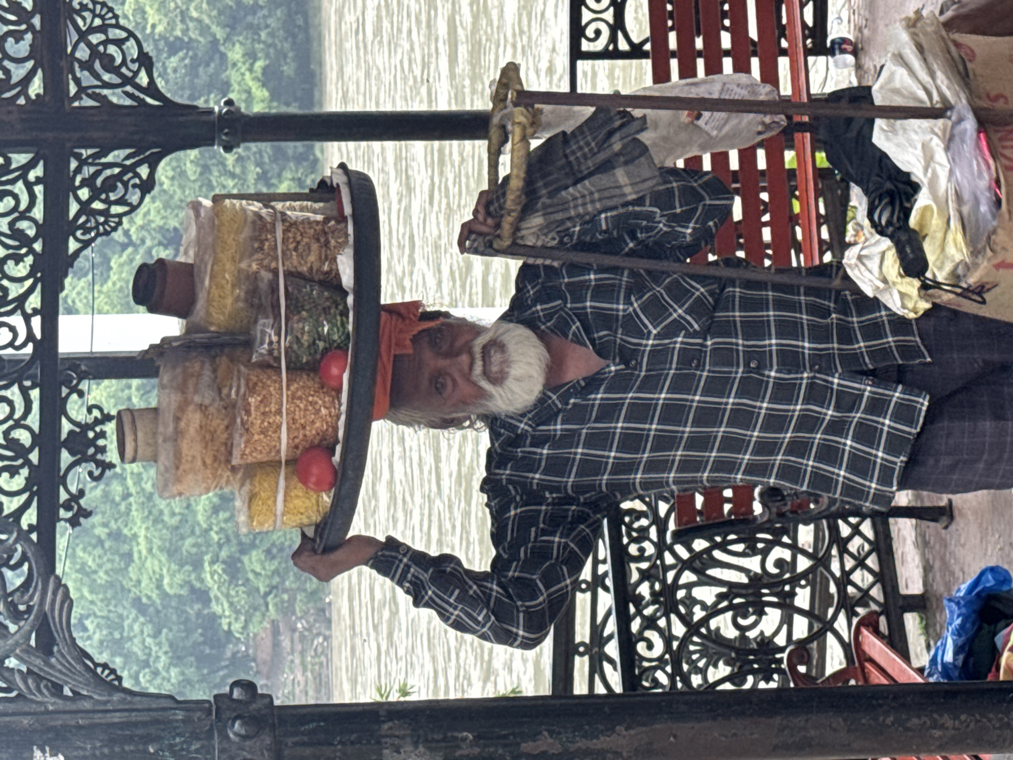 An elderly Indian man with a white beard and orange head covering stands in what appears to be a covered promenade by the Ganges River in Rishikesh, India. He is balancing a large, round tray on his head, filled with various snacks in plastic bags, including what looks like puffed rice and lentils. Two red tomatoes are also visible on the tray. He is wearing a blue and white checkered shirt and dark trousers. In his right hand, he holds a metal rod with some cloth draped over it, which is part of a cart he uses to sell his goods. The background shows the wide, rippling expanse of the Ganges River, with lush green trees on the opposite bank. Red benches are visible in the foreground, and ornate metal railings line the promenade. The lighting suggests an overcast day.