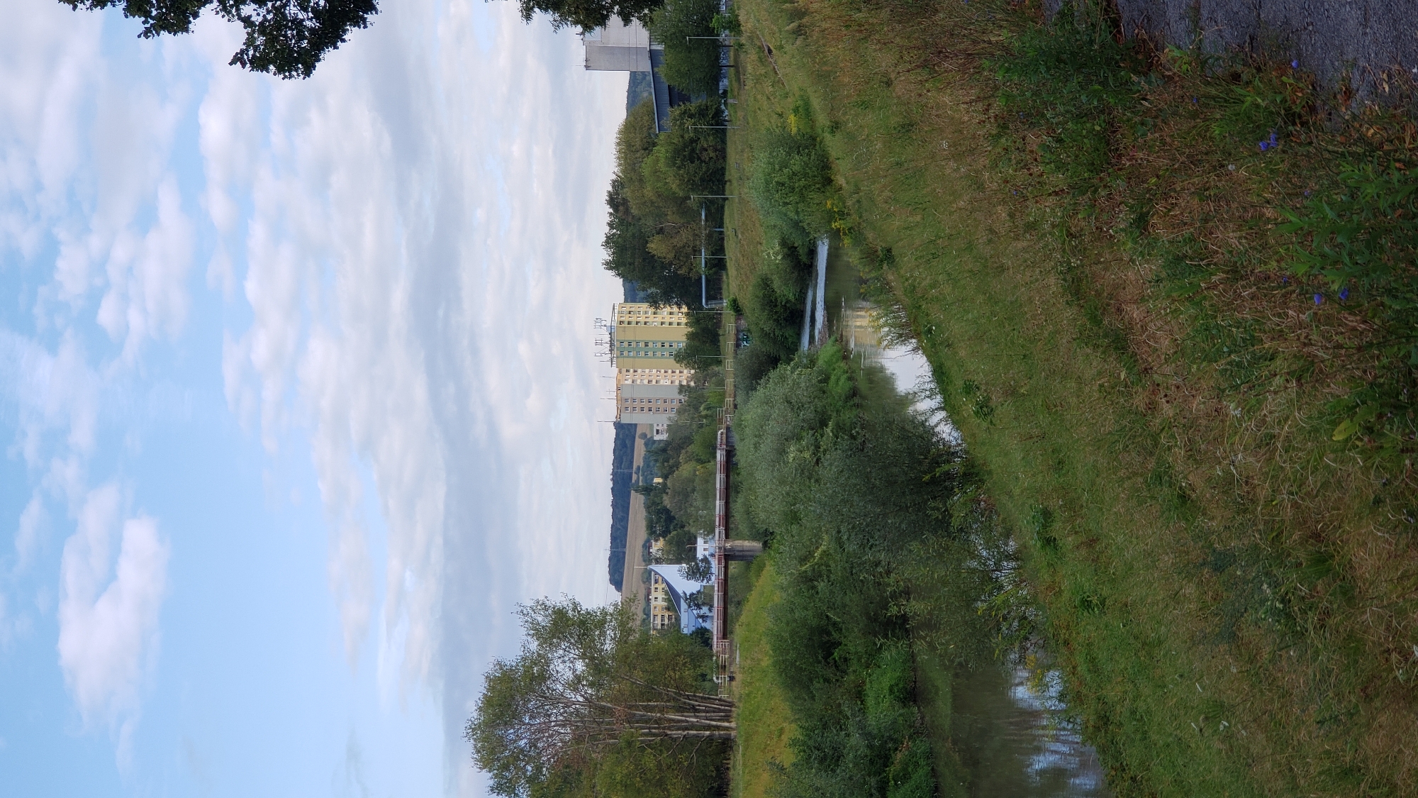 This image depicts a calm river flowing through a city, with a bridge spanning across it. On the left bank, there is lush green vegetation and trees, with a few tall, slender trees in the foreground. A multi-story residential building, painted in pastel colors, stands prominently in the background. To the right of the river, the bank is covered with grass and some smaller shrubs. The sky is partly cloudy, with a soft blue hue. The scene appears to be captured during daylight, possibly in the late afternoon or early morning given the lighting. The overall impression is one of a serene urban landscape with a strong natural element. This scene is likely located in Prešov, Slovakia, given the architectural style of the buildings and the presence of the river.