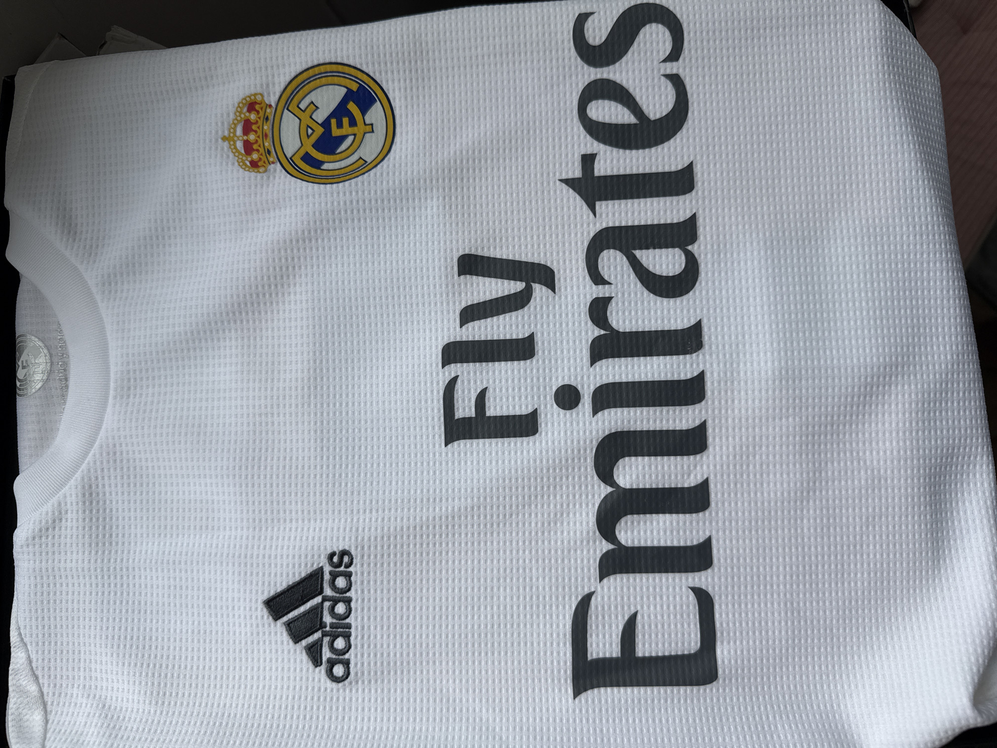 The image shows a white Real Madrid soccer jersey. The jersey features the Real Madrid crest on the upper left chest, the Adidas logo on the lower left, and the sponsor "Fly Emirates" vertically printed on the right side.