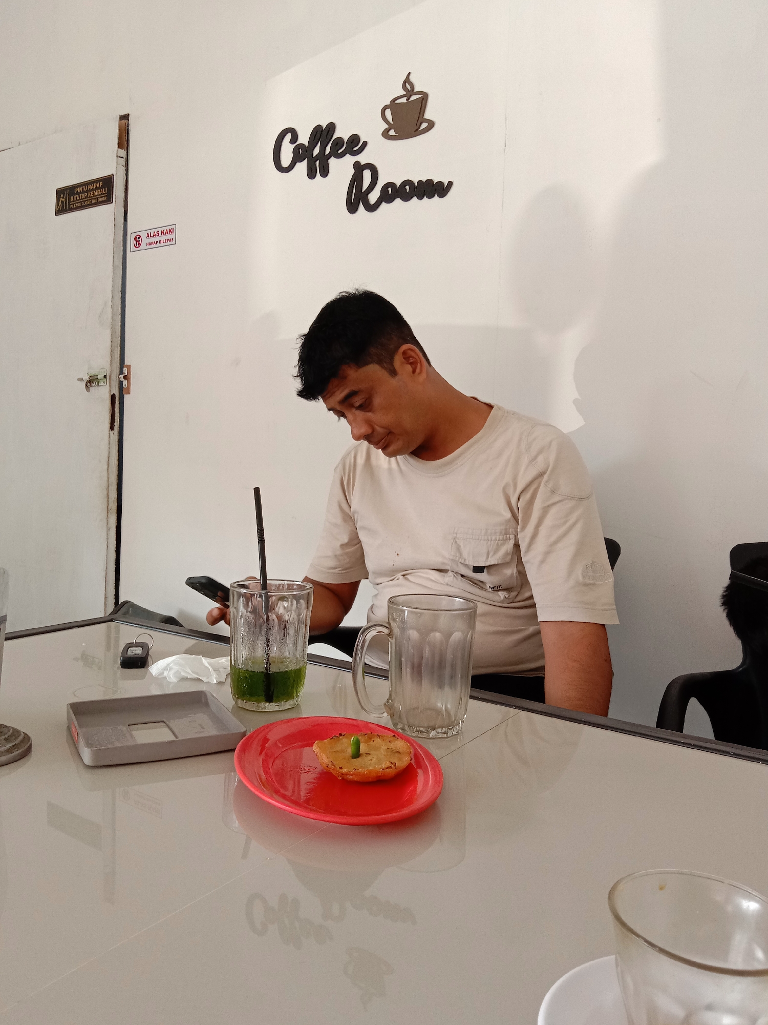 The image shows a man in a beige t-shirt sitting at a table in a coffee shop, likely in Lhokseumawe, Indonesia. He is looking at a cell phone in his hand. On the table, there is a glass of green beverage with a straw, an empty glass mug, a red plate with a snack topped with a green chili, a gray tray, a small black object (possibly a car key), and used tissues. In the background, the wall is white with the words "Coffee Room" written in a decorative font and a cup with steam, as well as Indonesian signs on a nearby door. The setting is a simple interior with a tiled table. The coffee shop has signage indicating rules or instructions, such as "Please Close The Door" and "Slippers off".