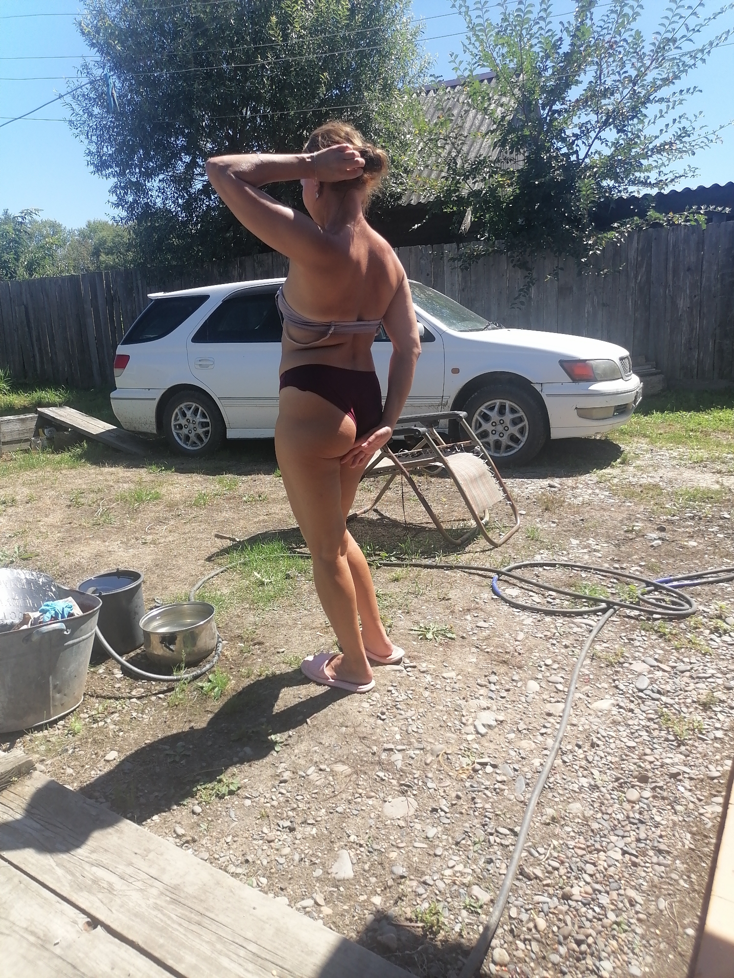 A woman, facing away from the camera, stands in a yard with a white station wagon parked behind her. She is wearing a dark-colored bikini bottom and a light-colored bra. Her hair is pulled up into a bun, and she is touching the back of her head. In the foreground, to the left, are three buckets: one large metal bucket on the far left containing what appears to be laundry and blue gloves, a black bucket next to it, and a metal pot filled with water to the right of that. A green garden hose is coiled on the ground to the right, near a collapsed metal lawn chair. The ground is covered with dirt, gravel, and scattered grass. A wooden fence runs along the back of the yard, with trees and a roof visible behind it. The sky is clear and blue. The scene is set in the city of Novopokrovka, Russia.