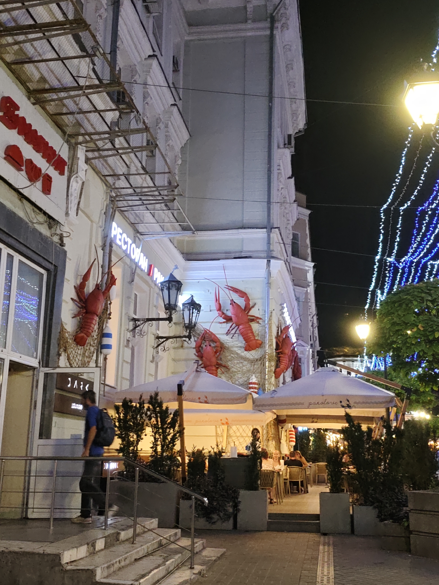 The image depicts a street scene at night in Rostov-on-Don, Russia. A restaurant facade is visible on the left, adorned with large red lobster decorations and netting. A sign above reads "РЕСТОРАН". On the right, an outdoor seating area for a restaurant is set up under umbrellas, with patrons seated at tables. The street is illuminated by streetlights and strings of blue and white fairy lights. A man wearing a blue shirt and backpack is seen walking up a set of concrete stairs. The architecture suggests an urban environment with older buildings.