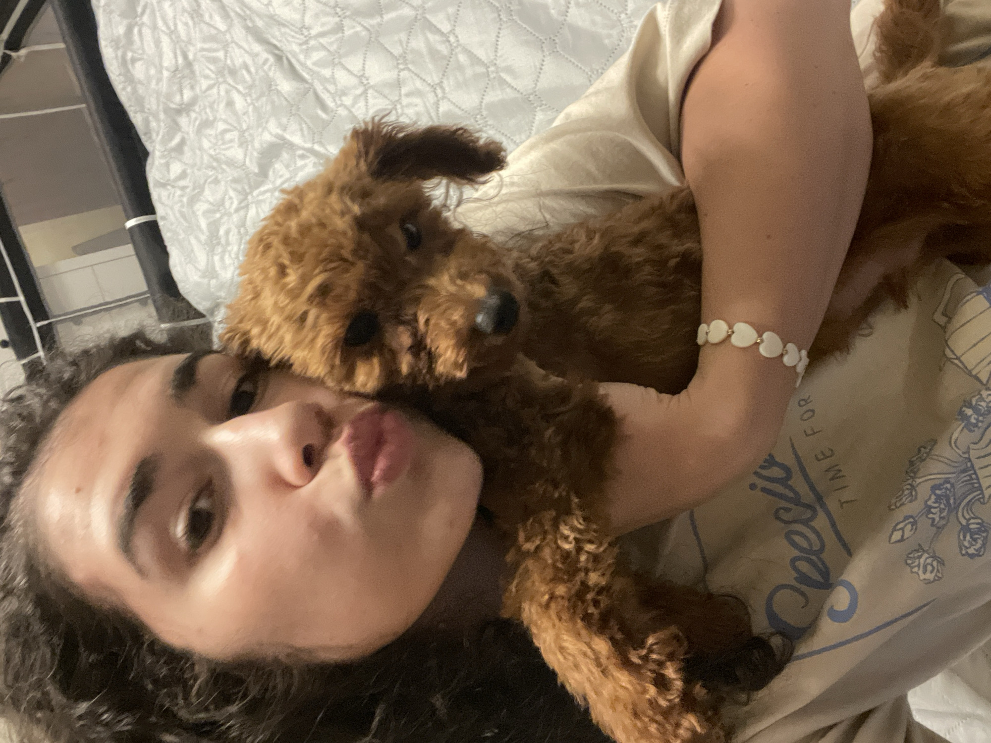 A person with curly dark hair is lying down on a bed with a light-colored quilted bedspread, holding a brown poodle. The person is wearing a light-colored t-shirt with a blue design and a bracelet with white heart-shaped beads on their wrist. They are looking at the camera with pursed lips. The location is indeterminate, as the image provides no visual clues indicating it is in Sofia, Bulgaria or any other specific location.