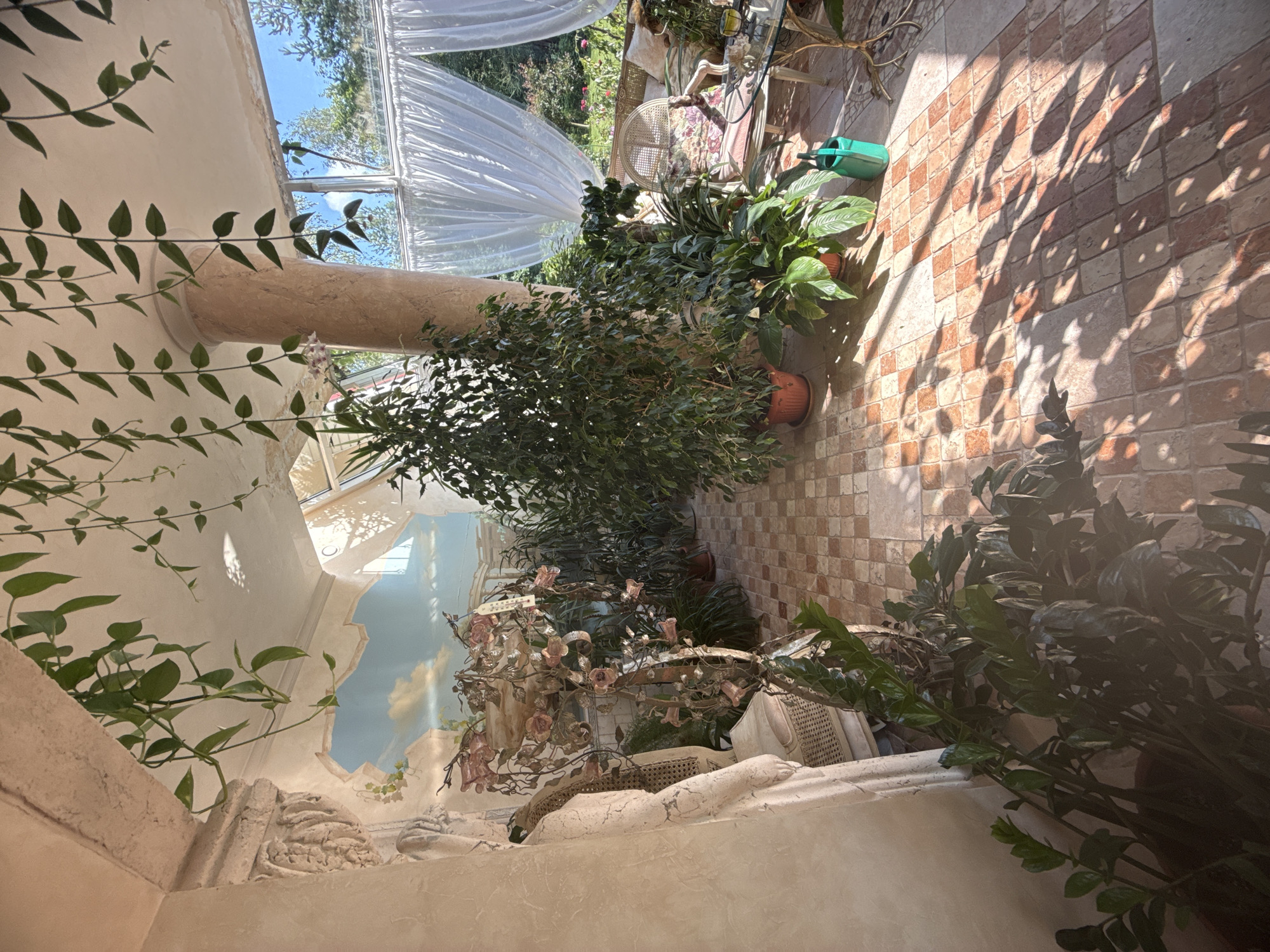The image depicts an indoor conservatory or sunroom with a tiled floor and a pale, textured ceiling. A large, leafy ficus tree stands prominently in the center, flanked by other various potted plants of different sizes and leaf shapes. Hanging vines with small green leaves cascade from the ceiling and an overhead archway. The room is adorned with classical architectural elements, including a fluted column and a sculpted relief on the wall. A mural of a sky with clouds is visible on the upper part of the wall. Natural light streams in from large windows draped with sheer white curtains, revealing a glimpse of green trees and foliage outside. In the foreground, a potted plant with dark, waxy leaves is partially visible. A wicker chair and a small glass-topped table with decorative metal legs are situated in the background to the right. A green watering can sits on the floor near a potted plant. The overall impression is one of a well-maintained, light-filled space filled with greenery. The location is stated to be in Yukki, Russia.