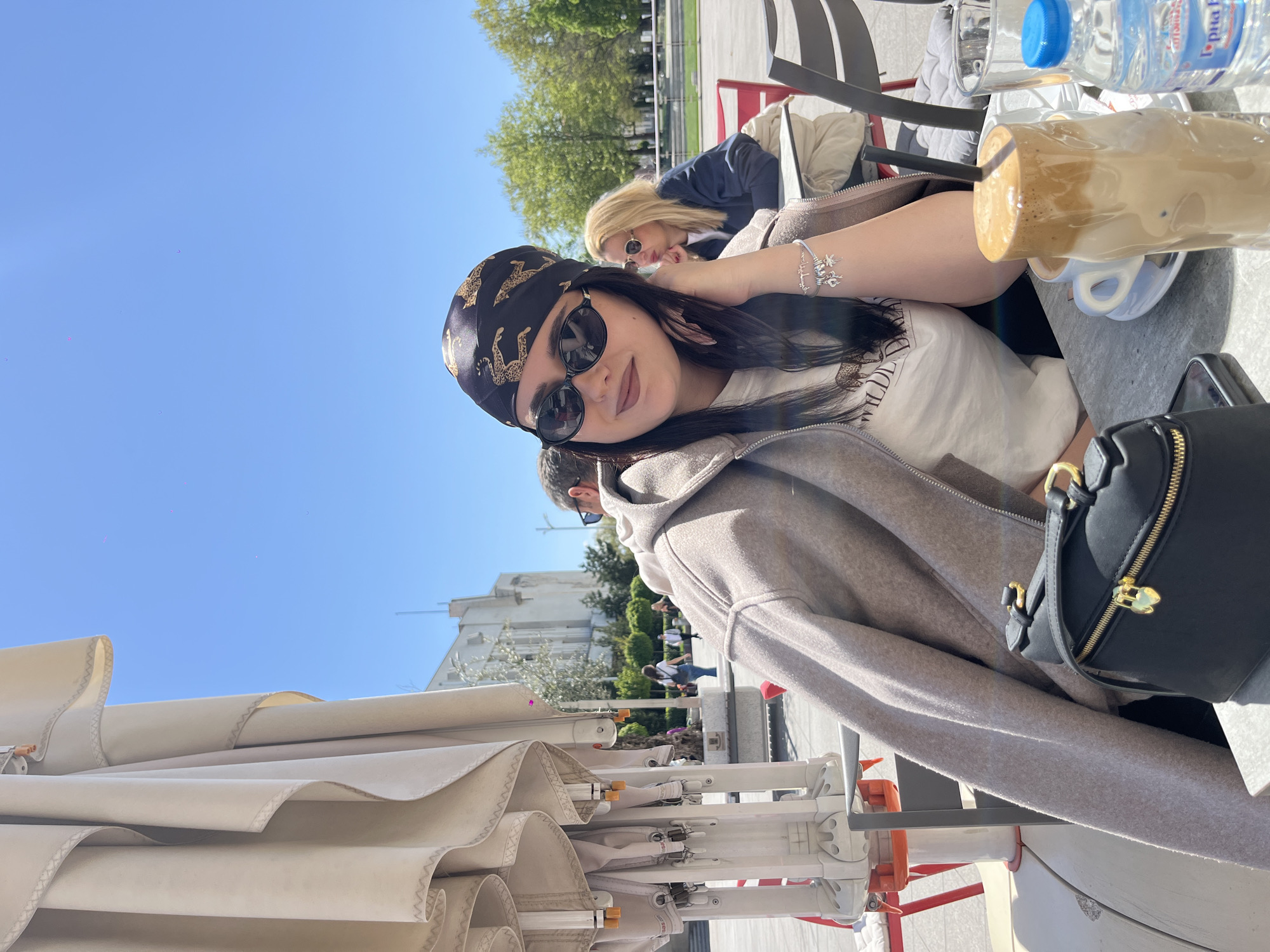 The image shows a young woman at an outdoor cafe in Plovdiv, Bulgaria. She is wearing sunglasses and a headscarf, a white t-shirt and a grey coat draped over her shoulders. She is seated at a table with a coffee drink and a bottle of water, a purse next to her. In the background, other people are at other tables. The sky is bright blue and buildings are visible in the distance. An open umbrella awning provides shade.