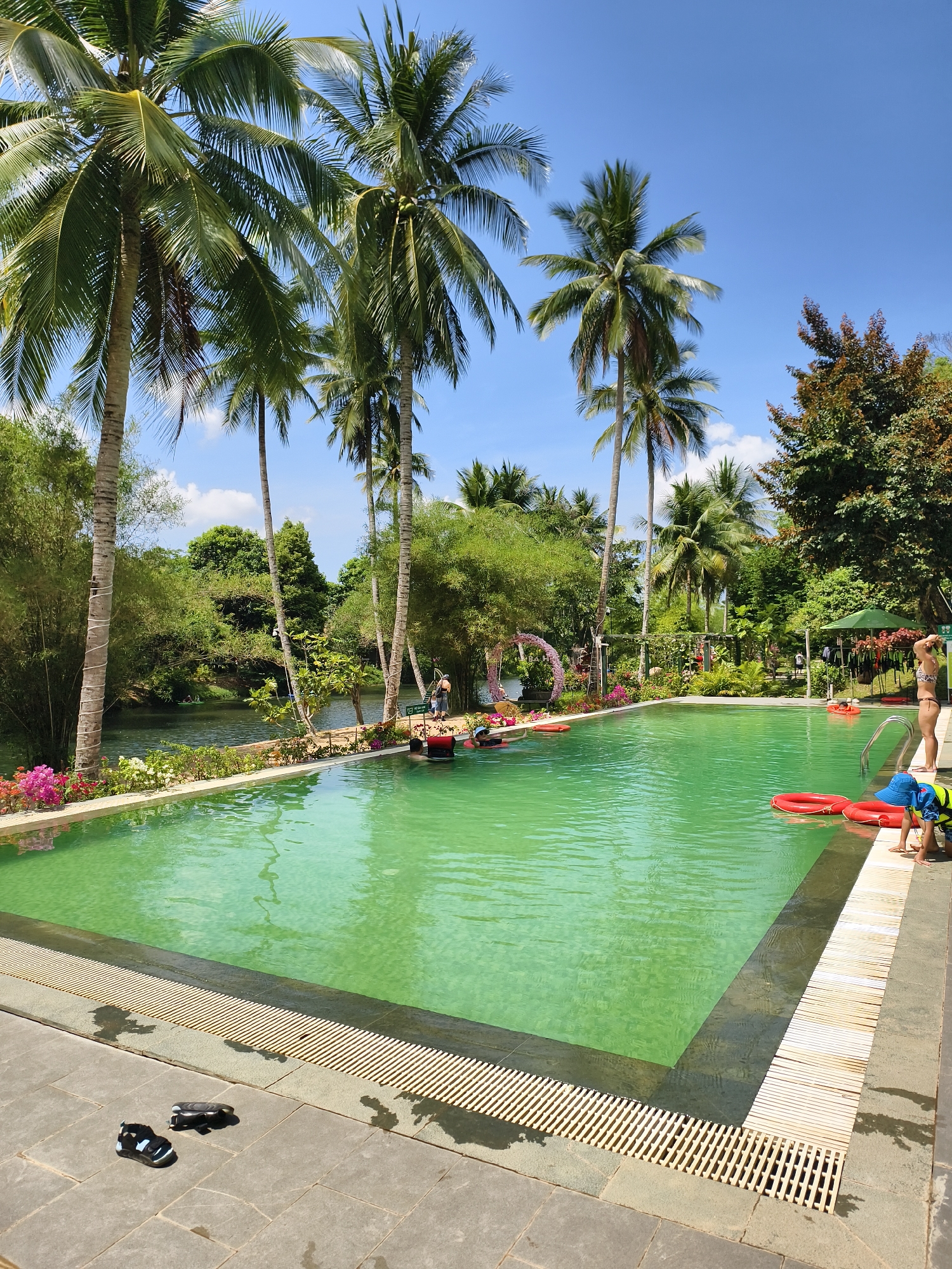 The image depicts an outdoor swimming pool adjacent to a river or stream, likely a resort or recreational area. The location is Unknown, Unknown.
The pool water has a greenish tint, possibly due to algae or mineral content. Palm trees line the pool's edge, and other foliage surrounds the area. Several people are present in and around the pool. Some people are swimming or floating with red inflatable tubes. A child wearing a blue hat and life jacket is near the pool's edge. A woman in a bikini stands near a ladder. There are children and some adults in the background near the water.
Near the pool's edge, a pair of sandals are visible on the tiled pavement. The pool has a drainage system with horizontal grills surrounding it. In the background, there is a floral archway and a bridge over the river. Other structures or possibly picnic areas are discernible in the distance. The sky is blue with scattered clouds, indicating a sunny day.