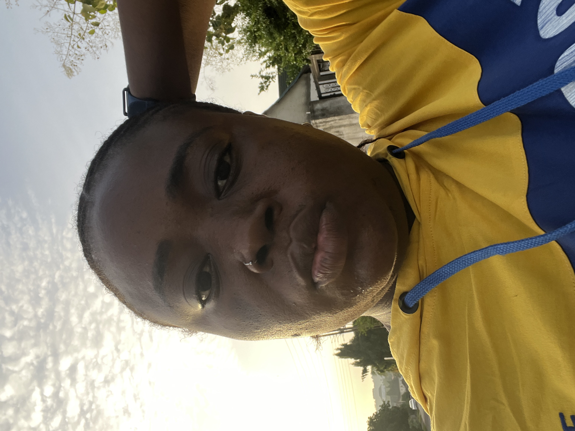 The image is a close-up selfie of a woman outdoors. She has dark skin, short dark hair, and a nose piercing. She is wearing a yellow and blue hoodie with a blue drawstring. She is also wearing a smartwatch. The background shows a sky with thin clouds, trees, and what appears to be a building in the distance. Based on the landscape and architecture, the image may have been taken in Abuja, Nigeria.