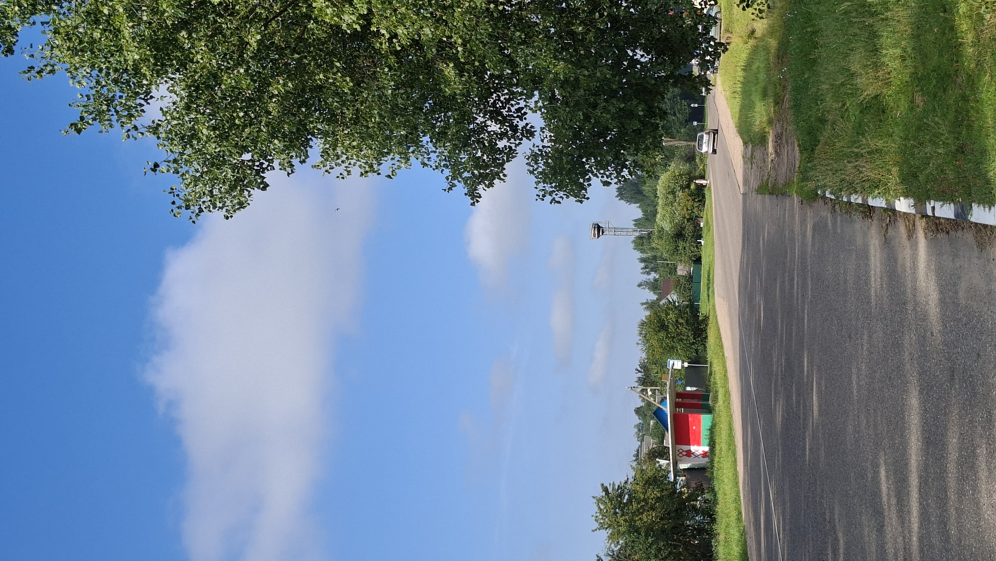 This is a vertical, eye-level, medium shot of a road in Забалоцкі сельскі Савет, Belarus. On the left side of the road is a bus stop shelter painted with red, green, and white designs. A silver car is driving away on the road towards the right. A large tree with green leaves dominates the right side of the frame. The sky is blue with scattered white clouds. In the distance, a water tower and several houses are visible amongst trees. The road has a white line marking the lane. The right side of the road has a grass verge with a concrete curb painted with black and white stripes.