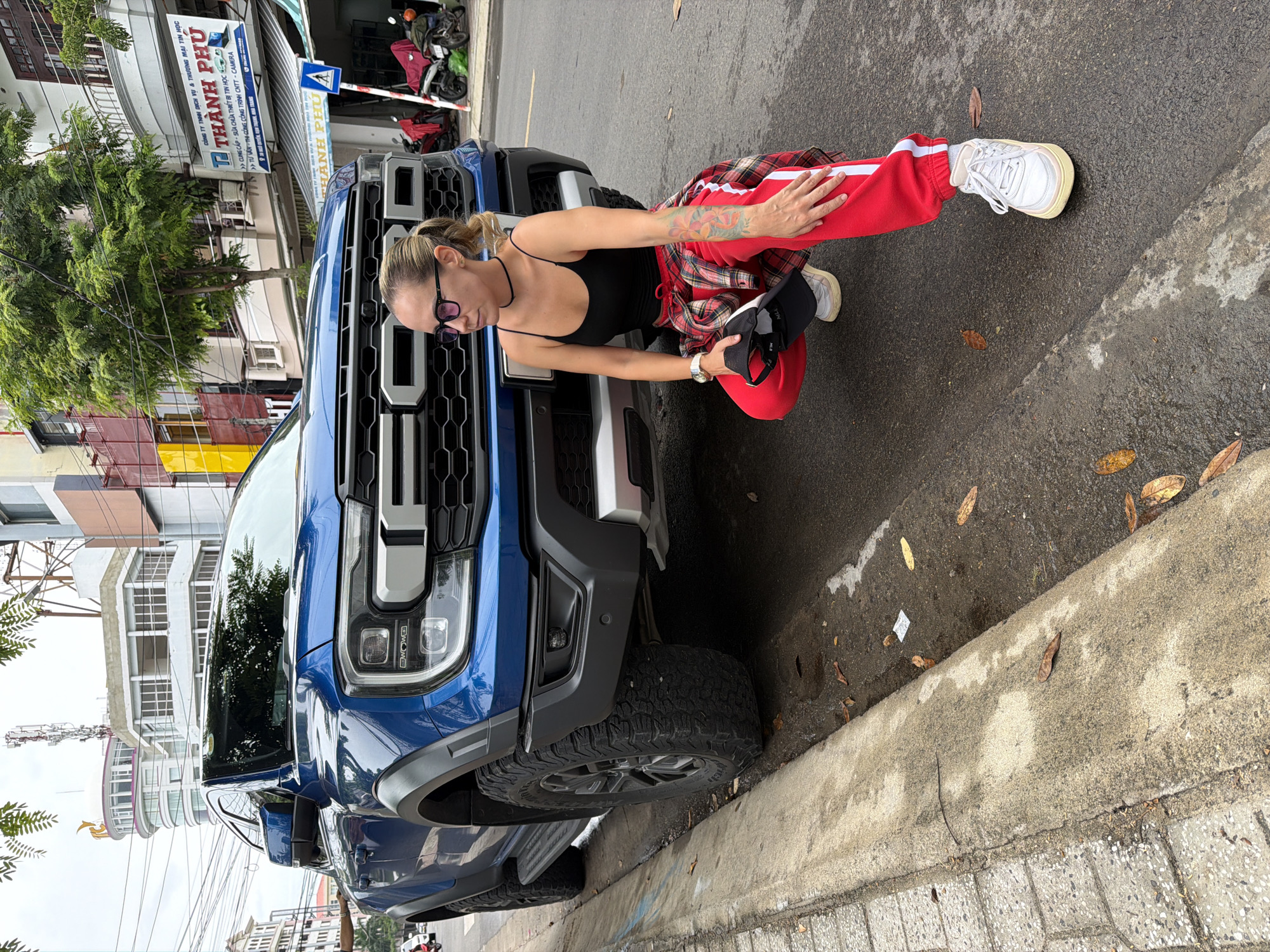 A woman with blonde hair pulled into a ponytail and wearing a black tank top, red track pants with white stripes, and white sneakers is crouching next to a blue Ford Ranger Raptor pickup truck parked on the side of a street in Nha Trang, Vietnam. She is wearing glasses and a choker necklace, and has a tattoo on her left arm. A red and black plaid shirt is tied around her waist, and she holds a black baseball cap in her left hand. The truck's front end is prominently displayed, with its large grille and headlights visible. The street appears wet, suggesting recent rain. The background shows buildings, trees, and electrical wires, characteristic of a Vietnamese urban environment. A blue and white sign with Vietnamese text is visible on one of the buildings, and a pedestrian crossing sign is on the right side of the image. A few motorcycles are parked in the background.