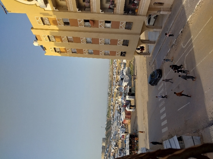 The image displays a detailed urban scene in Khan Yunis, Palestinian Territory. On the right, a multi-story residential building with a light tan facade and brown decorative window trim features numerous windows and balconies with white railings. A white water tank and solar panels are visible on its roof. Laundry hangs from several windows and balconies. The ground level incorporates open archways, with a white car parked beneath one. In the midground, a paved street contains a marked white pedestrian crosswalk. A group of approximately ten children are scattered across the street, engaged in play near a dark-colored sedan parked on the road. To the left of the street, several makeshift structures, constructed from tarps and corrugated metal, are present, with at least two adults standing in their vicinity. The background reveals an extensive, sprawling encampment composed of hundreds of white and grey tents and temporary shelters, stretching across a sandy landscape. In the far distance, large cylindrical tanks and other low-rise structures are visible against the horizon. The sky is clear and light blue, indicating natural daylight with shadows cast towards the left.