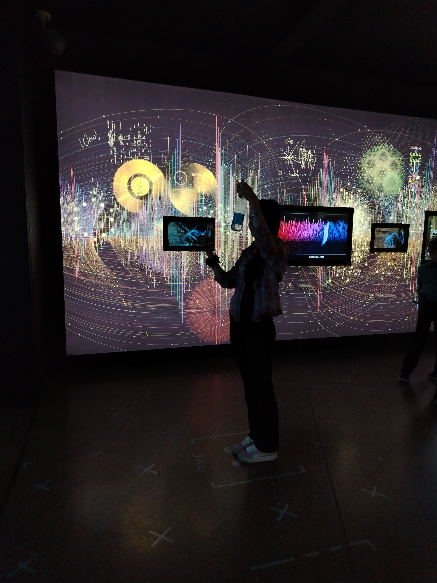 The image depicts an indoor exhibition space with a large, vibrant digital mural as the backdrop. The mural features an abstract design with swirling lines, geometric shapes, and what appear to be stylized representations of sound waves or data visualizations. Two smaller screens are mounted in front of the mural, displaying different visual content.
In the foreground, a person is silhouetted against the bright mural. They are holding up a smartphone or tablet, seemingly interacting with the exhibition by taking a picture or video. The person is wearing casual clothing, including a plaid shirt and dark pants, and light-colored sneakers. Their posture suggests engagement with the display. Further to the right, another person is partially visible, also in silhouette.
The floor of the exhibition space has white markings, possibly indicating interactive zones or pathways for visitors. The lighting is dim, with the digital mural providing the primary illumination. The overall atmosphere appears to be one of exploration and discovery, typical of a museum or science center. There is text visible on one of the smaller screens: "Frequency (Hz)". The word "Wow!" is also written in script on the large mural. The context of Moscow, Russia, is provided, but no specific Moscow landmarks are identifiable within the image itself. The time of day is indeterminate due to the indoor setting.