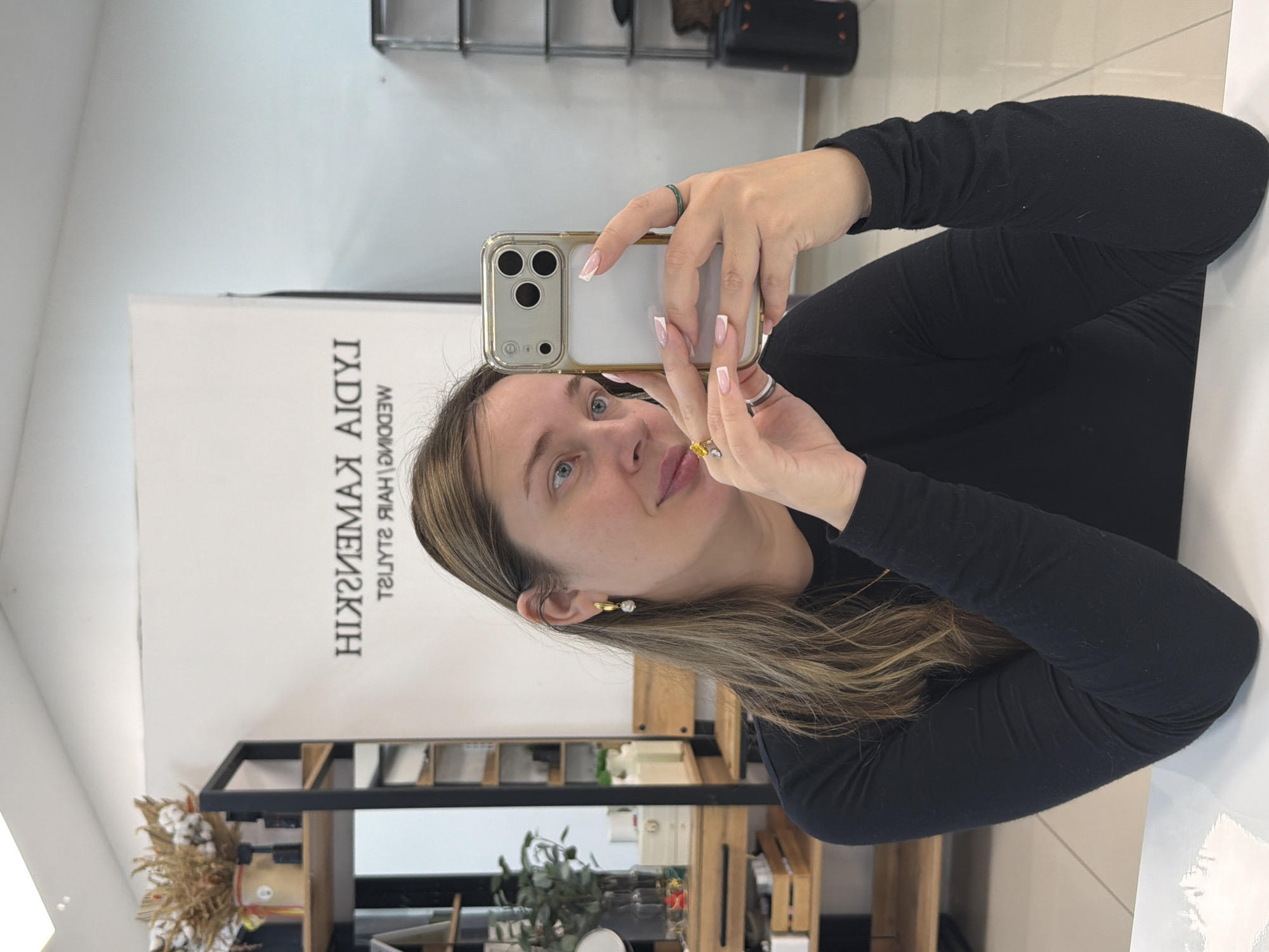 A single adult female is depicted taking a mirror selfie inside the "Lydia Kamenskaya Wedding Hair Stylist" salon, located in Chaykovsky, Perm Krai, Russia. The female, possessing long light-brown hair, blue eyes, and wearing a black long-sleeved top, holds a silver smartphone in a clear, gold-edged case with her left hand, angled to capture her reflection. Her right arm is bent, with her hand resting near her chin. She has a gold-toned earring with a white pearl-like drop in her visible left ear. Multiple rings are worn, including a silver-toned band on her left ring finger, a yellow-stone ring on her left index finger, and a green ring on her right ring finger. Her fingernails are manicured in a French style. The background features a white wall displaying the inverted text "LYDIA KAMENSKAYA WEDDING HAIR STYLIST". To the left, a wooden and black metal shelving unit contains items such as dried flowers in a pot, green plants in vases, and various white containers. To the right, a metal and clear-shelved unit is partially visible. The floor consists of light-colored tiles.