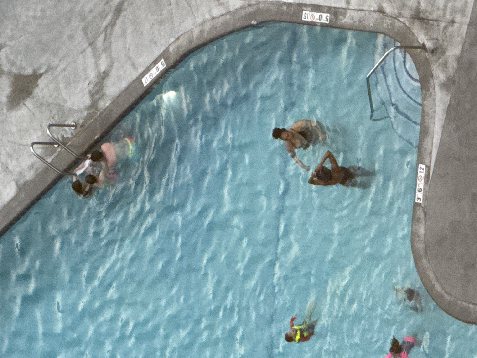 The image depicts a group of people swimming in a blue swimming pool. The pool has a concrete deck surrounding it. There are depth markers along the edge of the pool, reading "5 6 11", "5 0 15", and "3 6 12". Several individuals are visible in the water. In the upper part of the pool, a cluster of people are interacting, with some appearing to be children. To the left, a person wearing a yellow and red life vest is swimming. Towards the bottom right, two adult females are in the water, one with her hands on her head. A darker-skinned individual, likely a child, is also in the water near the bottom left. The overall scene suggests a recreational activity on a warm day. The image does not contain information about the specific location in Chicago, United States of America.
