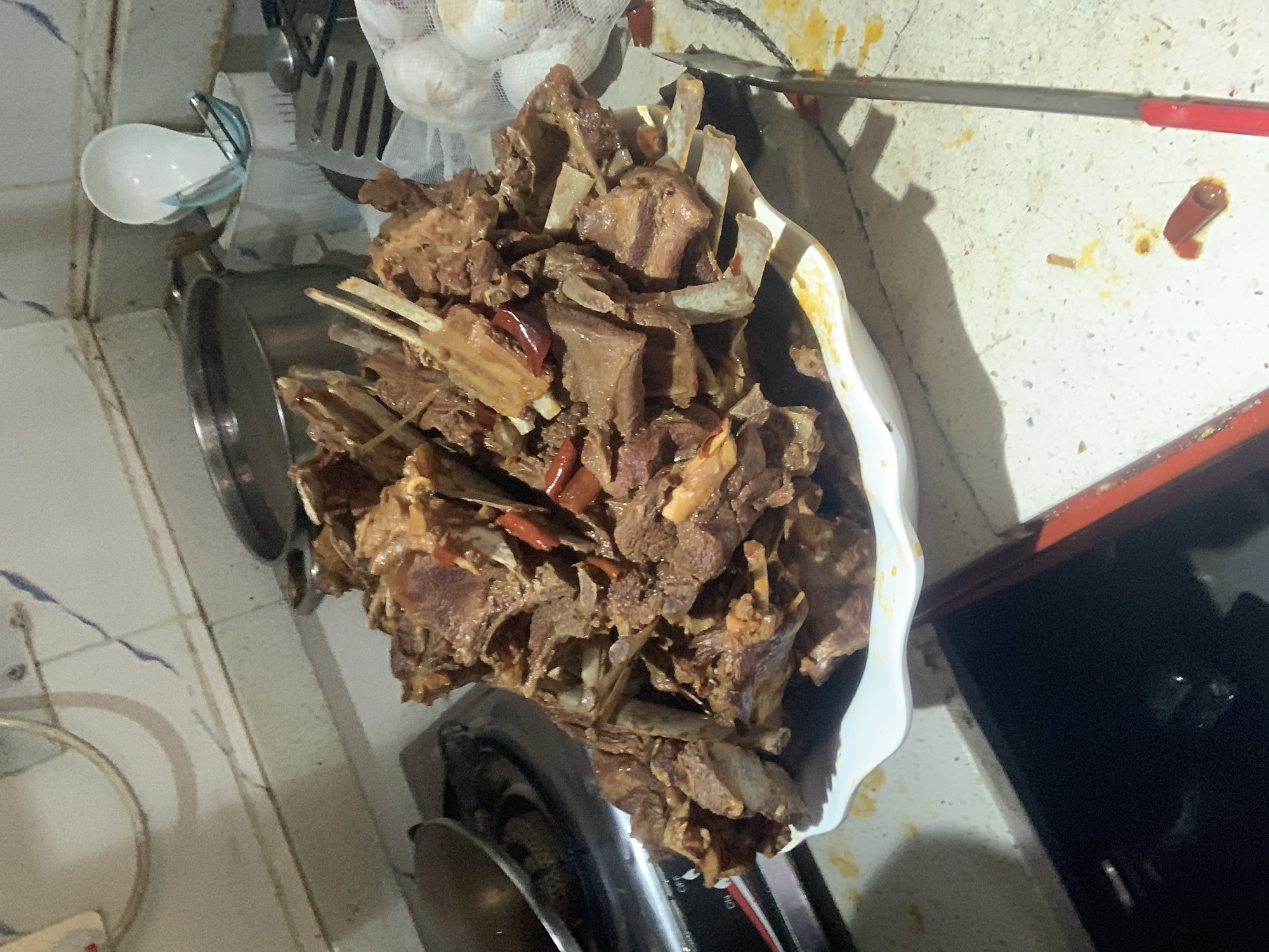 The image depicts a close-up, eye-level view of a large platter overflowing with a generous portion of braised meat, likely ribs, seasoned with what appear to be dried chili peppers. The meat is a rich brown color, indicating it has been cooked for an extended period, and some pieces show visible bone structure. The platter is white and has a slightly scalloped edge.
The platter of meat is positioned on a countertop in what appears to be a kitchen setting. To the left, a gas stove burner is partially visible, suggesting the food was recently prepared or is being kept warm. Behind the platter, various kitchen items can be seen, including a large metal pot, a white ceramic utensil holder with a white ladle and a blue handled spatula inside, and a stack of white mesh bags which might contain produce like onions or garlic. A metal spatula with a black handle lies to the right of the platter, and a long, slender metal rod with a red handle, possibly a cooking utensil or cleaning tool, is visible in the foreground.
The countertop itself is a speckled white material, and there are visible spills and stains of orange-yellow sauce, suggesting a slightly messy cooking or serving process. The background wall is tiled with white, square tiles, with a few blue lines forming a pattern. The lighting in the scene appears to be artificial and somewhat dim, creating shadows and highlighting the texture of the meat.
The image does not provide any specific information or visual cues to indicate a location within the city of Abuja, Nigeria. The scene is a generic kitchen environment focused on the food.