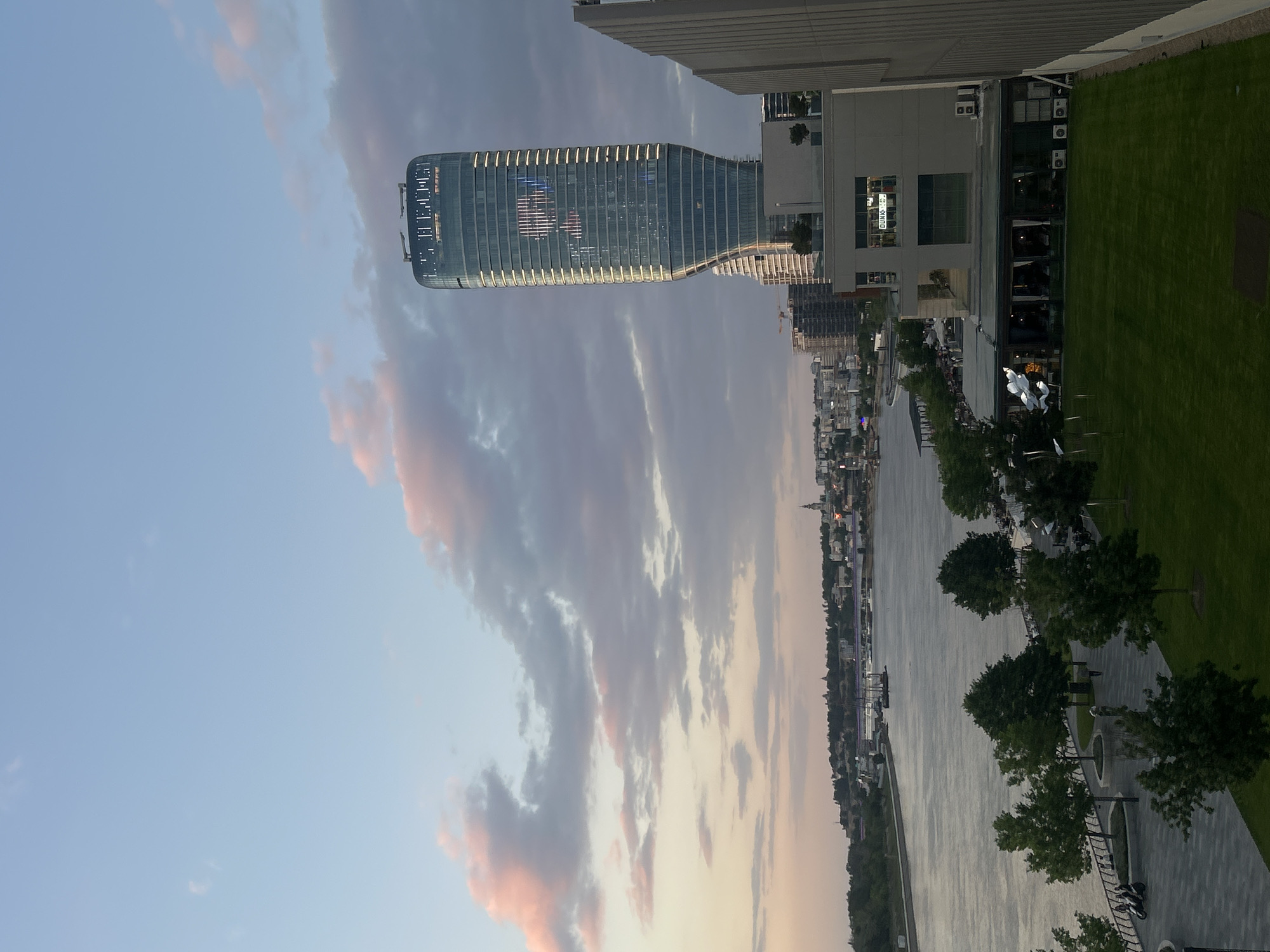 The image shows a view of Belgrade, Serbia, at dusk. The Danube River flows through the center of the frame, bordered by a park on the right with a paved walkway and trees. In the background, a skyline of buildings and structures can be seen along the riverbank. On the left, a modern glass skyscraper stands prominently against the sky, which is filled with clouds tinged with pink and orange hues. Adjacent to the skyscraper on the right is a building with a lawn and what appears to be a restaurant or cafe with outdoor seating.