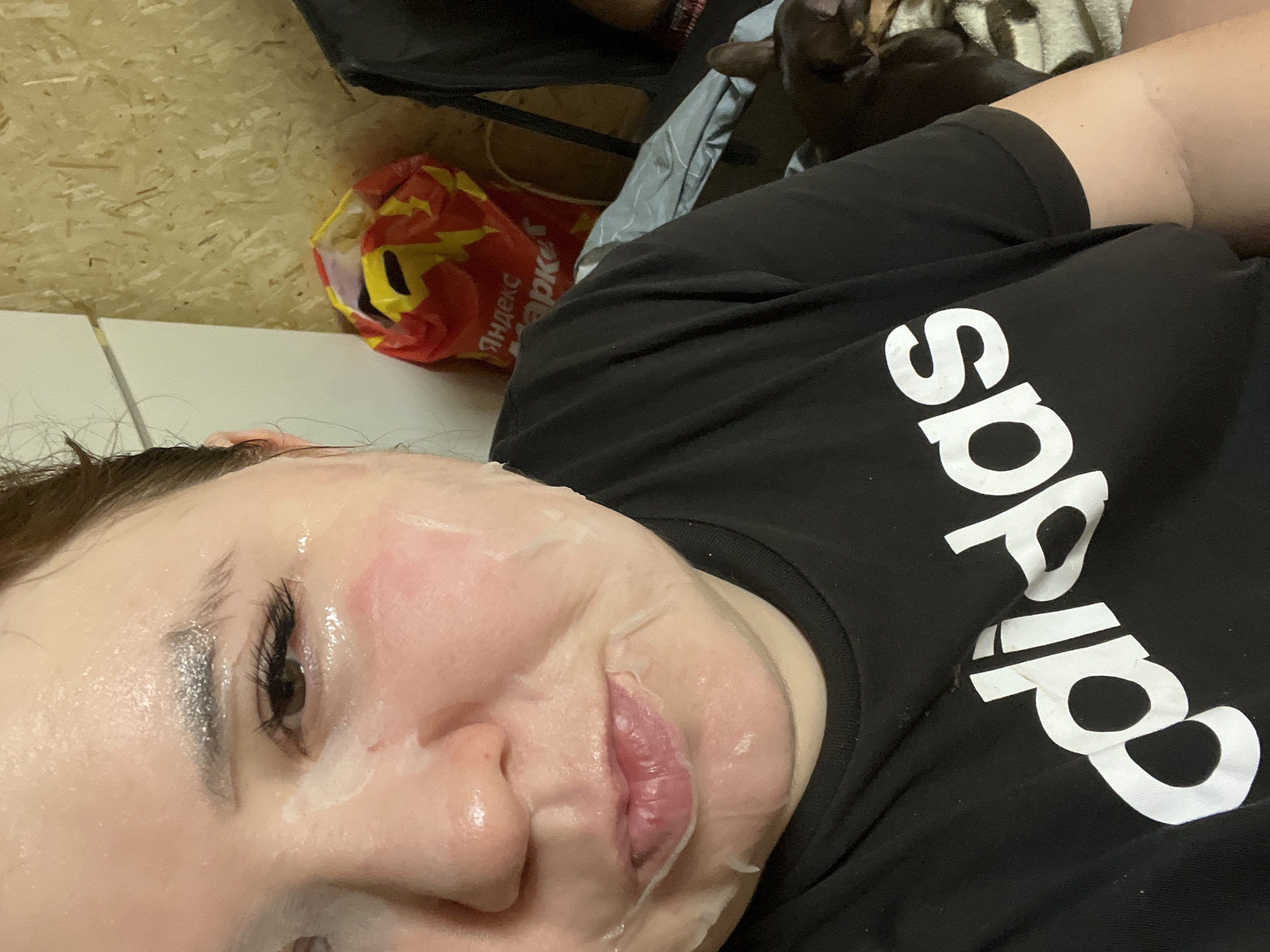 A woman is lying down wearing a facial mask. She is wearing a black Adidas t-shirt. A bag with Russian text on it is visible in the background. A black cat is in the upper right corner. The walls are wood paneling. The location is Chaikovsky, Russia.