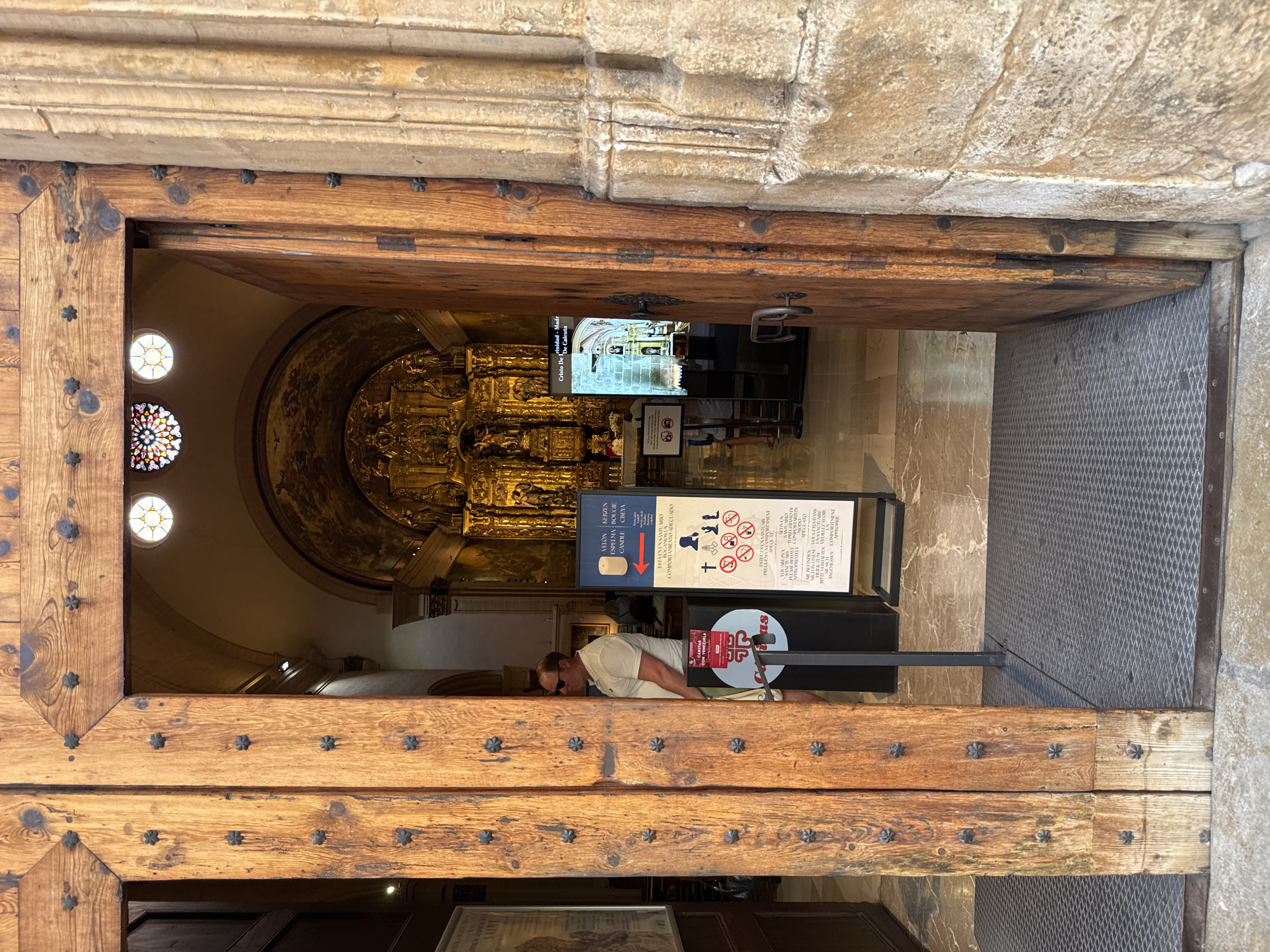The image shows a view through a large, weathered wooden double door, opening into the interior of a cathedral. The cathedral's ornate golden altar and stained-glass windows are visible in the background. To the right of the doorway, a man wearing sunglasses and a light-colored polo shirt is partially visible, looking into the cathedral. A digital information screen stands to the left of the doorway, displaying an image of a crucifix and text in Spanish. A sign with pictograms indicating appropriate behavior within a sacred space is also present. The floor inside the cathedral is made of marble tiles. The doorway itself is constructed from thick wooden planks, reinforced with metal studs, and leads from a stone exterior to the cathedral's interior.