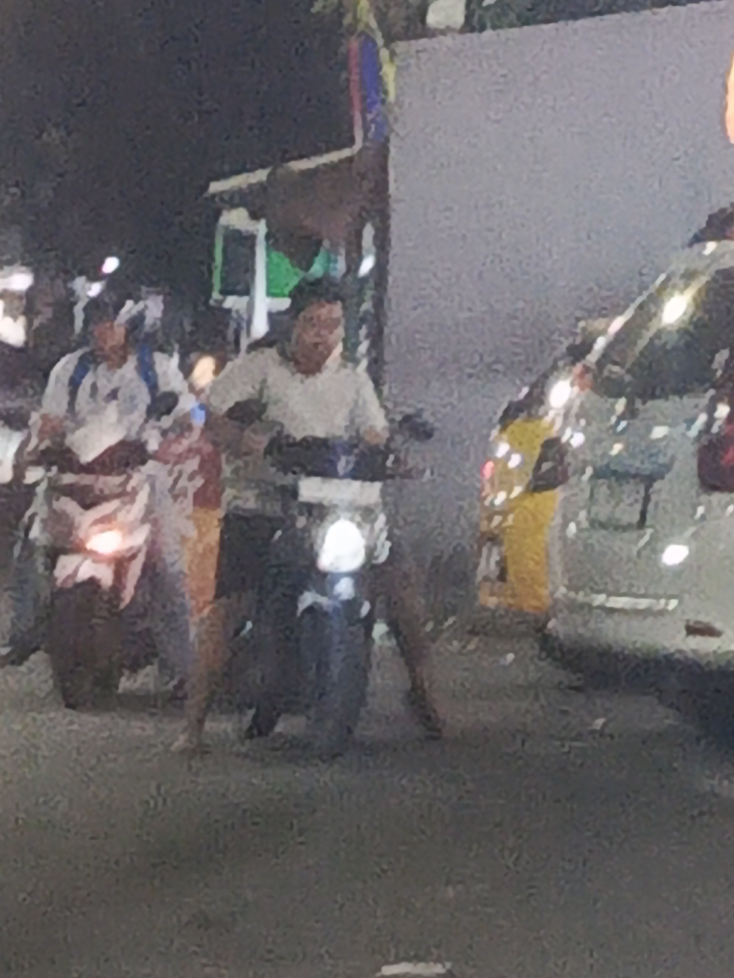 The image depicts a nighttime street scene in Surabaya, Indonesia, with two individuals on motorcycles. The person on the left is wearing a light-colored shirt and a backpack, riding a white scooter with a prominent headlight. The person on the right is wearing a light-colored t-shirt and appears to be riding a darker-colored motorcycle with a visible headlight. The background shows a dimly lit street with what appears to be buildings and a white vehicle, possibly a van or car, to the right. The ground surface is paved, suggesting a road or parking area. The lighting is low, creating a grainy and somewhat blurred image typical of nighttime photography.