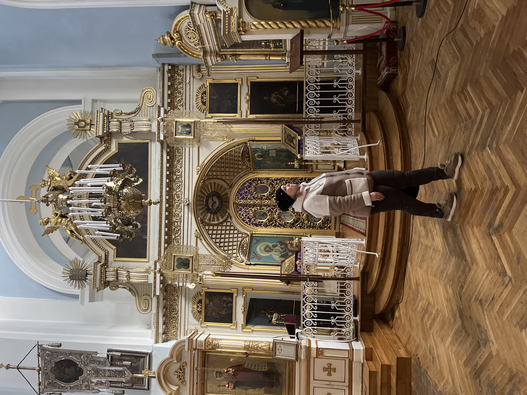 The image displays the interior of the Transfiguration Cathedral (Spaso-Preobrazhensky Cathedral) in Saint Petersburg, Russia. An adult female is positioned in the lower-right mid-ground, walking or standing with her head turned left, looking upwards towards the main altar area. She has light skin, dark hair visible beneath a black beanie, and is wearing a two-tone beige and light brown zippered jacket with a fur/shearling-like collar and trim, dark trousers, and light grey sneakers with a visible fur lining.
The background is dominated by a multi-tiered, highly ornate iconostasis, primarily white and gold, featuring intricate carvings, moldings, and numerous framed icons depicting saints and biblical scenes. The central Royal Doors are purple with gilded grilles and circular icons. To their left and right are deacon doors and additional icons. A large, gilded, multi-armed chandelier with candle-like lights and angelic figures hangs from the ceiling in front of the iconostasis. The upper walls are painted a light blue, accented by white arches and architectural elements. A large, ornate silver processional banner, featuring an icon, hangs on the upper left wall.
The foreground consists of a light-toned wooden parquet floor laid in a herringbone pattern, displaying some darker areas of apparent wear. Wooden steps lead up to a raised platform (solium), which is bordered by a white, ornate metal railing with scrollwork. A wooden lectern or stand is visible behind the railing on the right, with another similar stand to the left. Red velvet ropes strung between black stanchions restrict access on the far right. A black electrical cable traverses the floor from the right side. The scene is brightly lit, showcasing the intricate details of the ecclesiastical architecture and decor.