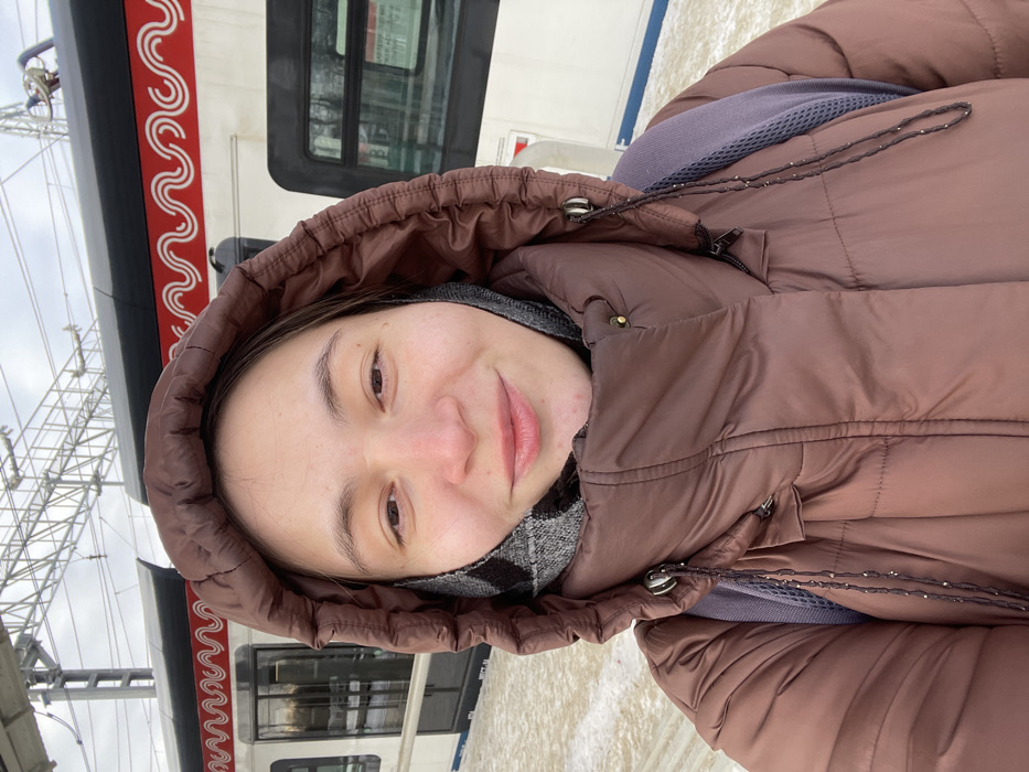 The image captures a young adult female taking a close-up selfie at a train station platform, identified as being in Fryazevo, Russia. The subject faces the camera with a slight smile, wearing a brown hooded puffer jacket and a gray and black patterned scarf or neck gaiter. A purple backpack strap is visible over her right shoulder. Behind her, a white train with a prominent red and white wavy "M" pattern, indicative of Moscow Central Diameters (MCD) rolling stock, is stationary at the platform. The platform itself is light-colored and exhibits traces of snow or ice. Overhead electrical lines and support structures are visible above the train against a cloudy sky. Bare trees are faintly discernible in the background beyond the railway.