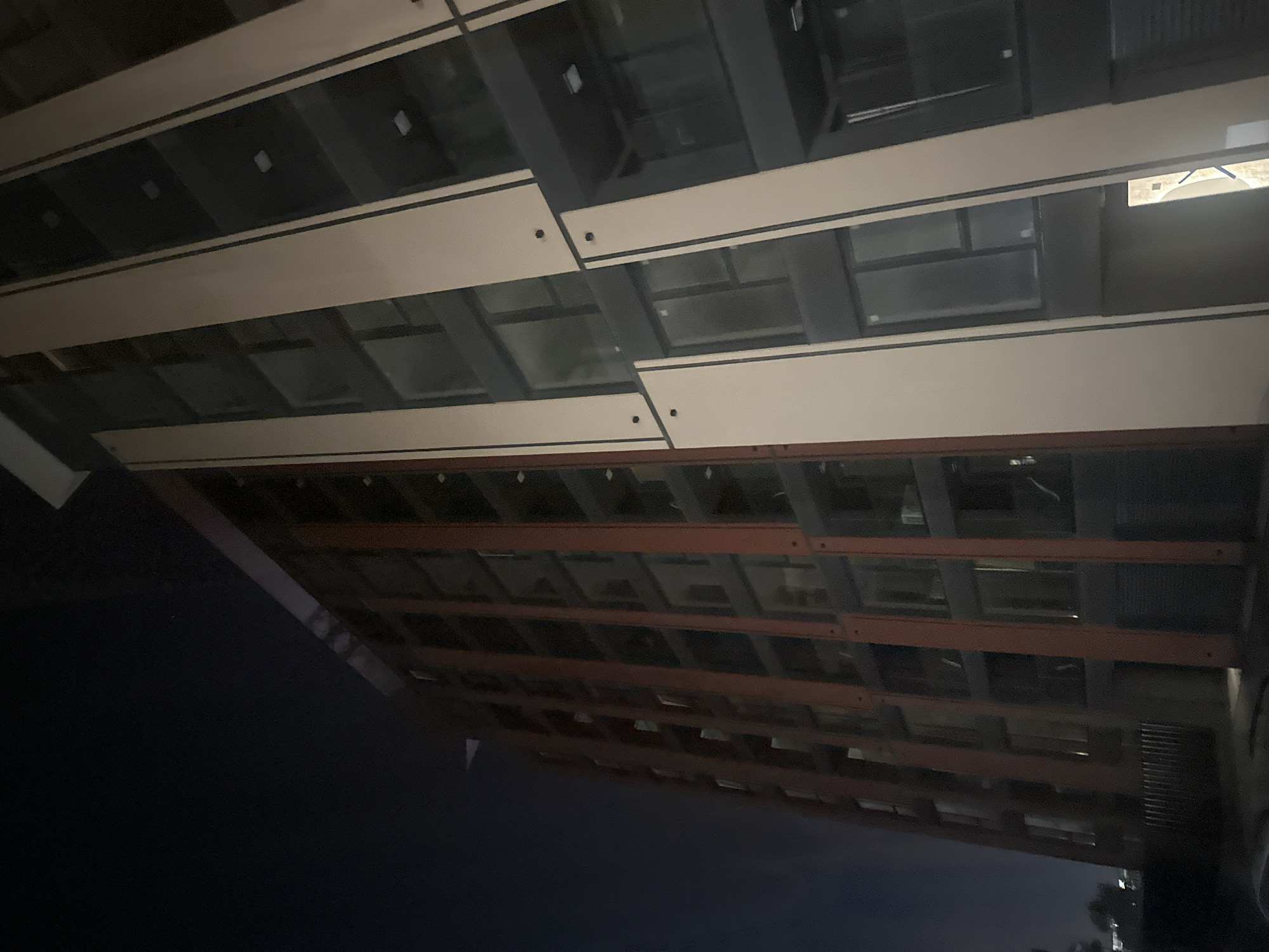 A nighttime, low-angle, full-shot perspective looks up at a tall, modern building under construction. The building has a dark, grey facade with wide, beige vertical beams and thin, red, vertical structural elements that frame recessed dark grey windows. The sky above is a deep, uniform black. A few faint white lights are visible within some of the windows. At the bottom left, a dark, shadowy area suggests ground level with a faint hint of foliage and a metal fence. The overall impression is one of an unfinished urban structure against a dark night sky.