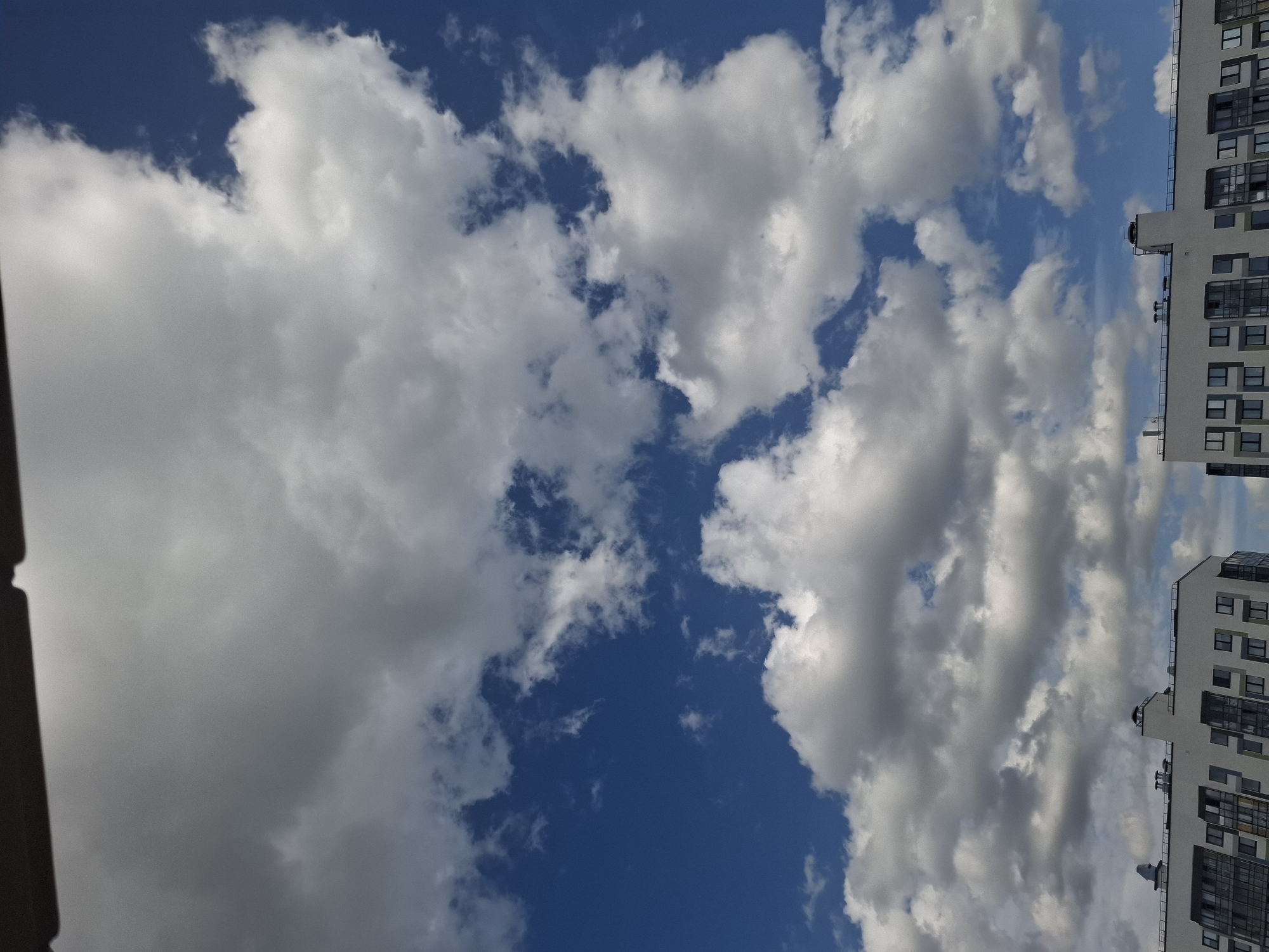 The image shows a view of the sky and the tops of modern residential buildings in Bugry, Russia. The sky is predominantly blue, filled with fluffy white and gray clouds, suggesting a partly cloudy day. The buildings are contemporary in design, with white facades, dark-framed windows, and balconies. Some buildings feature visible ventilation systems or chimneys on their rooftops, along with railing systems. The perspective is from a low angle, looking upwards. There are no people visible in the image.