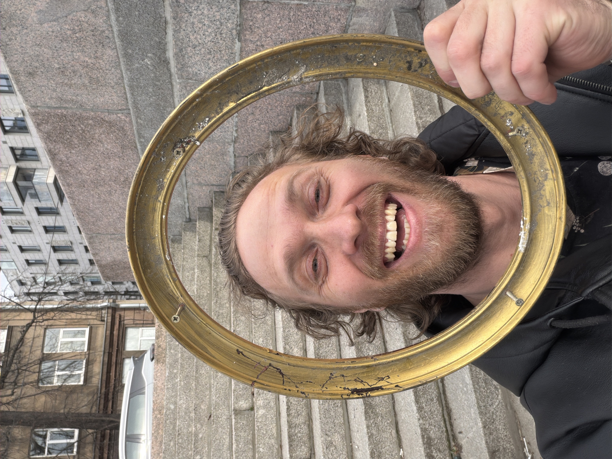 A man with shoulder-length curly hair and a beard smiles broadly at the camera while holding a gold-colored, circular frame around his head. The frame has minor damage and visible screws. The man wears a black jacket over a patterned shirt.
The setting appears to be outdoors, likely on or near a set of stone stairs. In the background, various buildings are visible, including one with repetitive window patterns. Based on architectural details, the location is highly likely to be Saint Petersburg, Russia. The presence of bare tree branches suggests the photo was taken during late autumn or early spring.