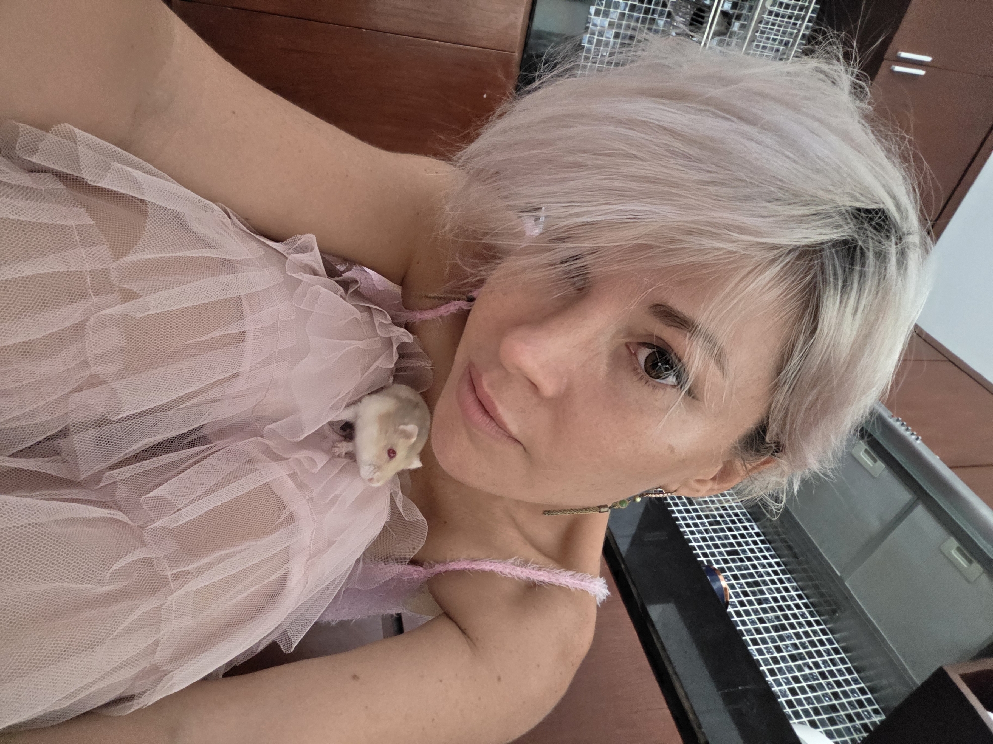 A woman with platinum blonde hair, wearing a pink lace dress, is holding a small, light-colored hamster. The hamster is resting on the woman's chest. The woman's right arm is visible, and her left arm is bent, supporting the hamster. She is looking directly at the camera with a neutral expression. The background appears to be an indoor setting, possibly a kitchen, with wooden cabinets and a stainless steel range hood. The image was taken in North Kuta, Indonesia.