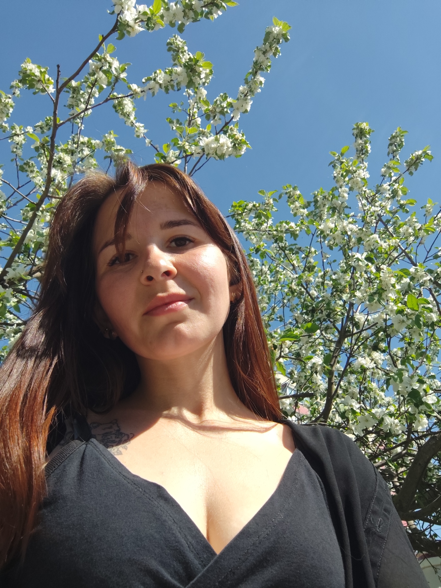 The image is a low-angle selfie of a woman with long, auburn hair and fair skin. She is positioned in front of a blossoming tree with white flowers set against a clear blue sky. The woman is wearing a black V-neck top. A partial view of a tattoo can be seen on her chest. The photo appears to have been taken outdoors in the city of Chaikovsky, Russia, as indicated by the prompt. The focus is on the woman's face, with the tree and sky serving as a backdrop. The sunlight casts shadows on her face, giving it a warm glow.