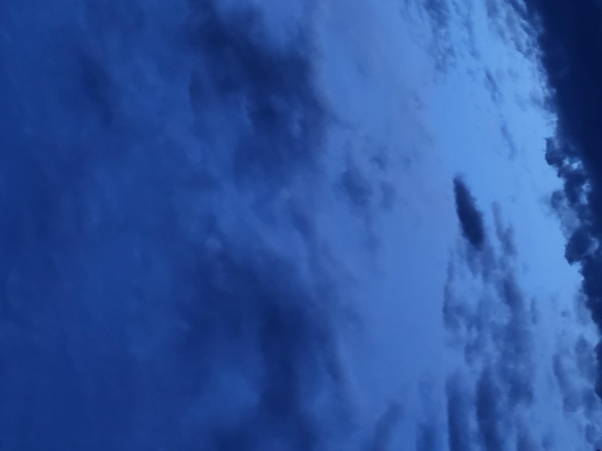 The image displays a dark blue sky with scattered clouds. The lighting suggests it is either early morning or late evening. No specific landmarks or identifiable features are visible to pinpoint a location within the city of Jelgava, Latvia. There are no people, subjects, objects, or actions depicted in the image. The environment is solely the sky and clouds.