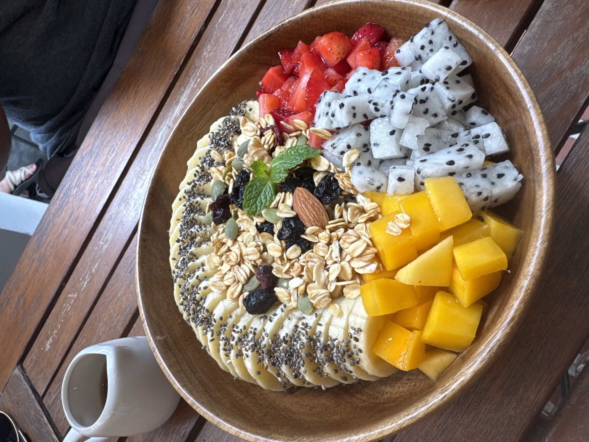 The image shows a wooden bowl filled with a colorful assortment of fruits, granola, and seeds, situated on a wooden surface. The fruit selection includes sliced bananas topped with chia seeds, diced mango, cubed dragon fruit, and diced strawberries. A mix of granola, nuts, and dried fruit is arranged centrally within the bowl, and a sprig of mint garnishes the arrangement. In the lower left corner of the image, a white ceramic cup sits adjacent to the bowl. The bowl is placed on a weathered wooden table with visible grain. A portion of a person's leg, wearing a sandal, is visible in the upper left background. The photograph appears to be taken in an outdoor setting in the city of Đà Nẵng, Vietnam, suggested by the style of wood furniture commonly found in Vietnamese outdoor cafes.