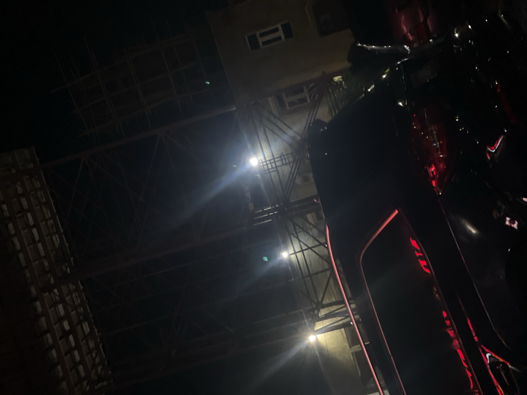 The image shows a nighttime scene in Lugbe, Nigeria, featuring a building under construction with exposed steel scaffolding and support structures. Lights illuminate the framework. A portion of a dark vehicle with red accent lighting is in the foreground.