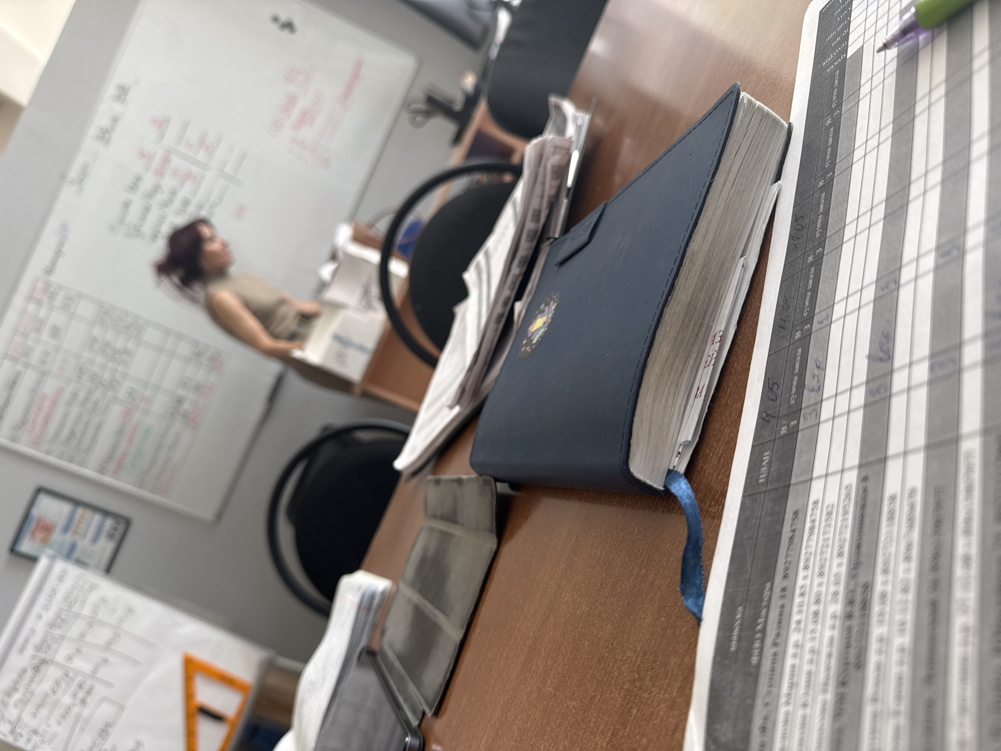 The image depicts an office scene with a blurred figure in the background, a white board with writing, and a desk covered with various items. On the desk, there is a blue notebook with a blue ribbon bookmark, sheets of paper with typed data (possibly financial records), and a pen. The text on the paper appears to be in Russian, including names, phone numbers, and potentially transaction details. The background figure seems to be sitting at a desk in the office. The location might be Tolyatti, Russia, based on the language on the paper.