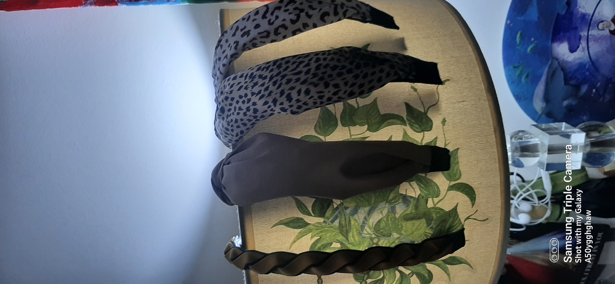 The image shows a close-up of three headbands resting on the top edge of a lampshade. The lampshade has a pattern of green leaves and vines. From left to right, the headbands are a dark brown braided band, a plain dark brown band, and a leopard print band. The background is a lit ceiling. Below the lampshade, part of a crystal lamp base and a decorative plate with a marine life scene are visible. A watermark in the bottom left corner reads "Samsung Triple Camera Shot with my Galaxy A50yggghaw". The image does not contain any information that would indicate its location in Dubai, United Arab Emirates.
