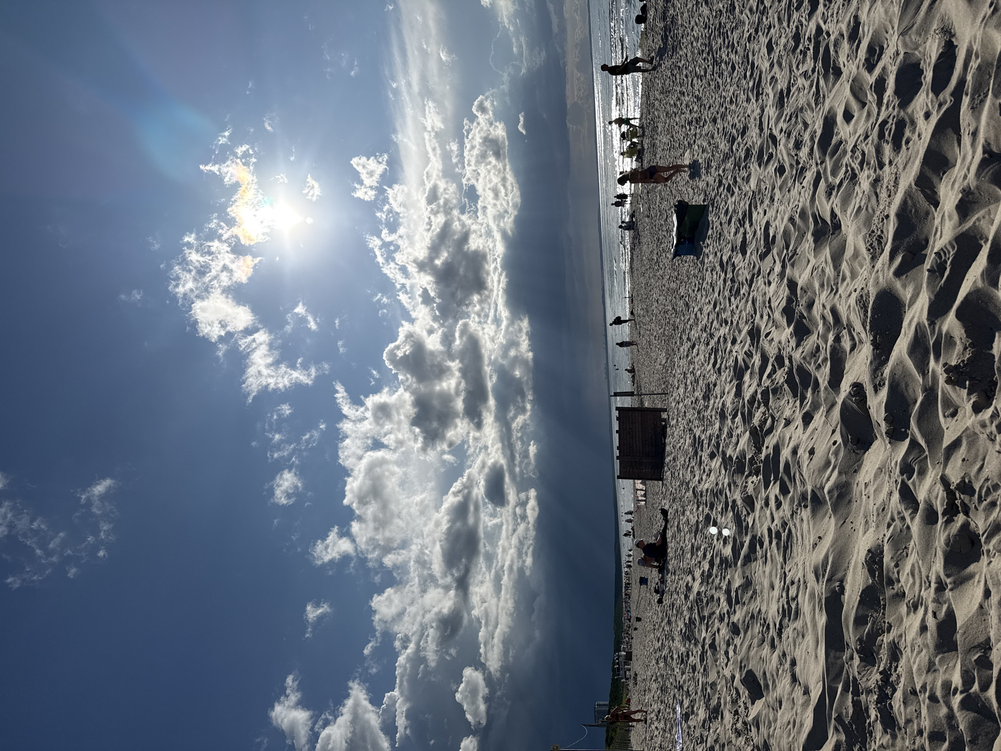 The image depicts a crowded beach scene on a sunny day in Międzyzdroje, Poland. The foreground is dominated by textured sand dunes, casting shadows under the bright sunlight. In the midground, numerous people are scattered across the beach, some sitting, others standing, and a few walking towards the water. A wooden structure, possibly a changing room or shelter, stands near the water's edge. The sea is visible in the background, with gentle waves lapping at the shore. The sky is a dramatic mix of blue and dark, heavy clouds, with the sun shining through, creating visible light rays. The overall impression is one of a lively summer day at the seaside.