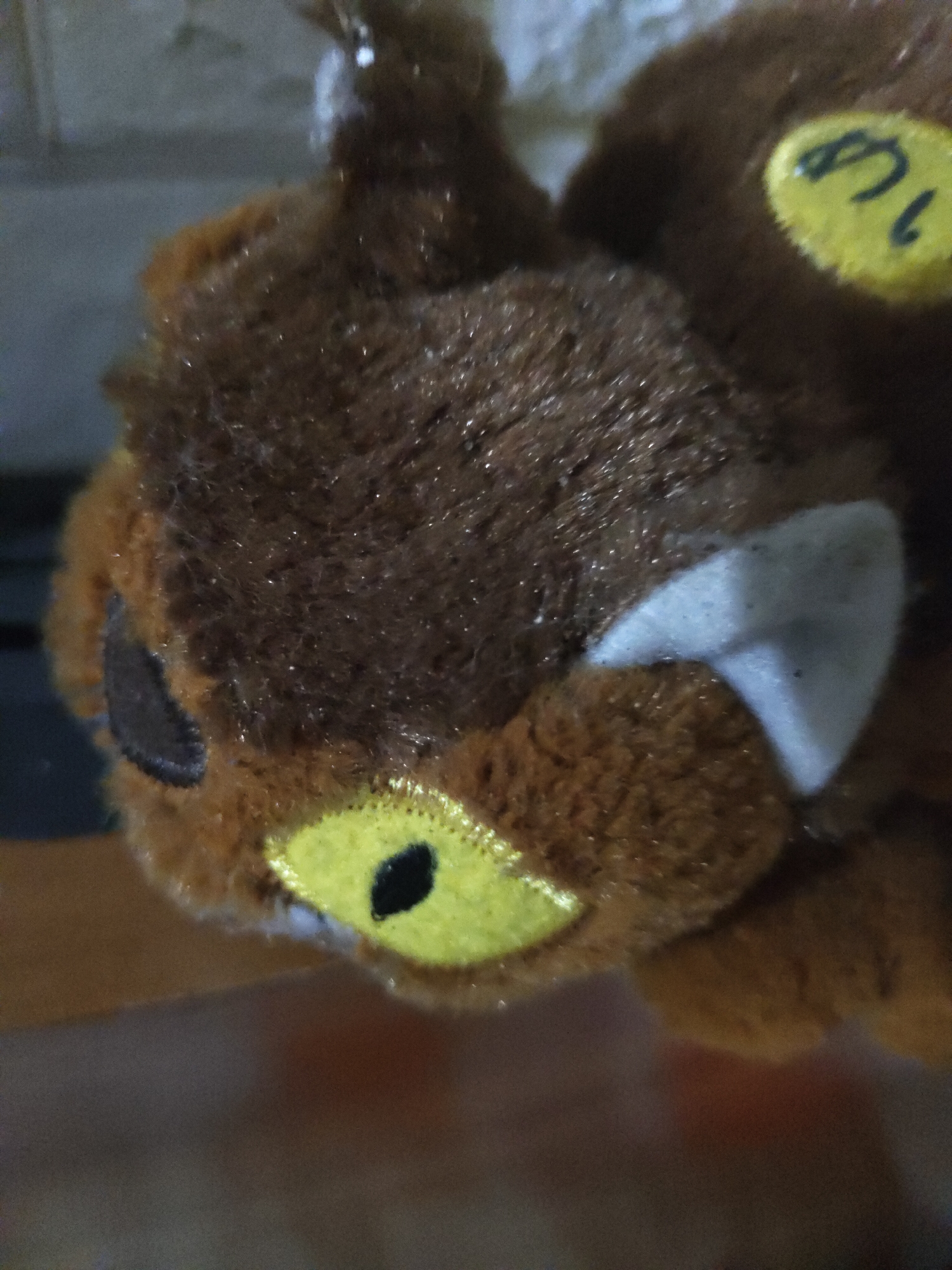 A close-up photograph captures a brown, furry plush toy with a dark brown head and lighter brown muzzle. The toy features two prominent yellow, oval-shaped eye patches, each containing a central black, vertically elongated pupil. On the upper right side of its head, a triangular white patch is visible, resembling an ear. On the opposing left side of its head, a smaller, dark gray, oval-shaped marking is present. The top-rear section of the head displays a yellow, irregular-shaped tag or marking with black markings resembling the number "8" and a partial "1" or "L". The fur exhibits a fine texture with numerous tiny reflective points, indicating light reflection. The background is out of focus, presenting a blurred gradient of light gray, white, and brown tones, suggesting an indoor environment. The image was captured in the city of Unknown, Unknown.