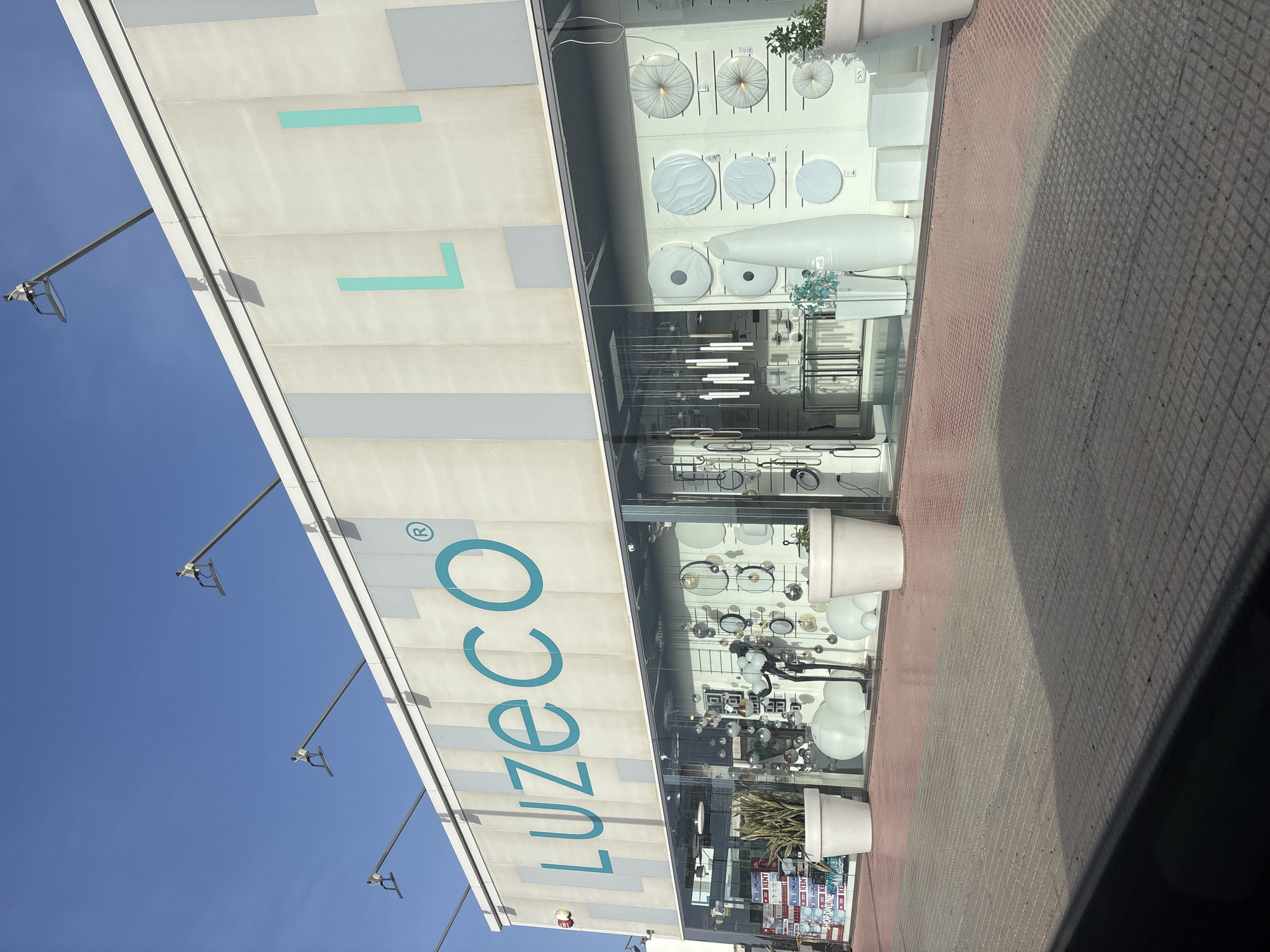 The image shows the exterior of a retail store named "LUZCO" located in Orihuela, Spain, specializing in lighting fixtures and home decor. The store features a large glass storefront displaying various modern lighting designs, decorative wall panels, and large white planters. Above the storefront, the "LUZCO" logo is prominently displayed in turquoise lettering against a light-colored facade with vertical paneling. The building has an industrial design with several external light fixtures extending from the roof. The ground in front of the store is paved with textured tiles and a pinkish walkway. The sky is clear blue, indicating daylight. No people are visible in the image.