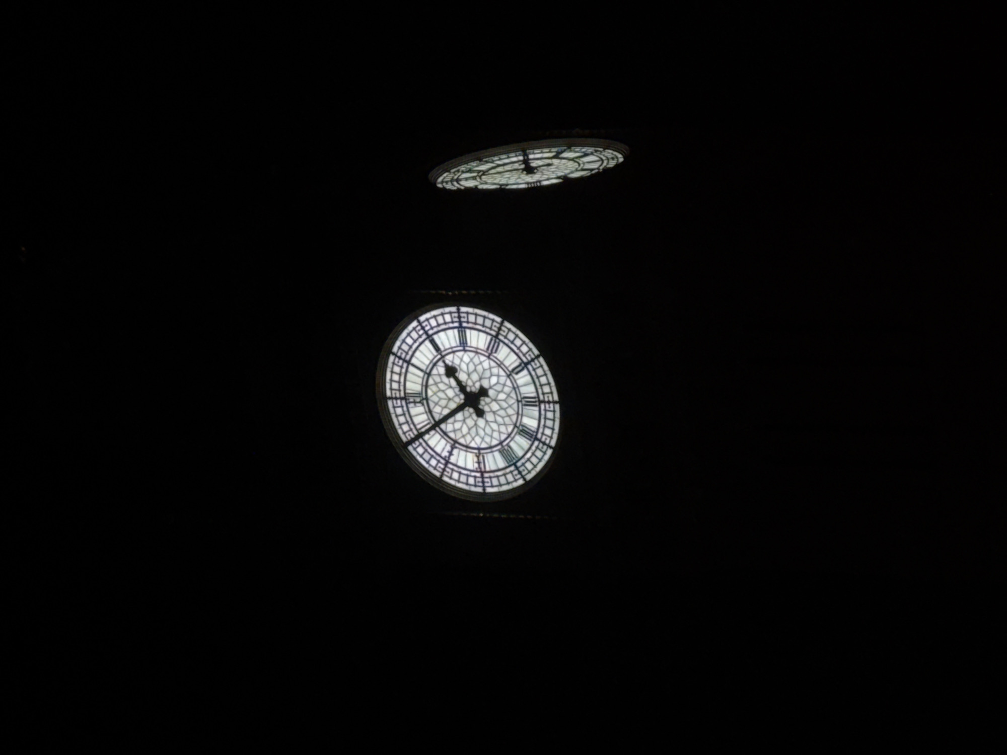At night, the illuminated clock faces of the Elizabeth Tower, commonly known as Big Ben, are visible against a dark sky in London, United Kingdom. Two faces are discernible: one is seen almost head-on, showing the clock's hands, while the other appears above it and at an angle. The surrounding environment is dark, emphasizing the brightness of the clock faces.