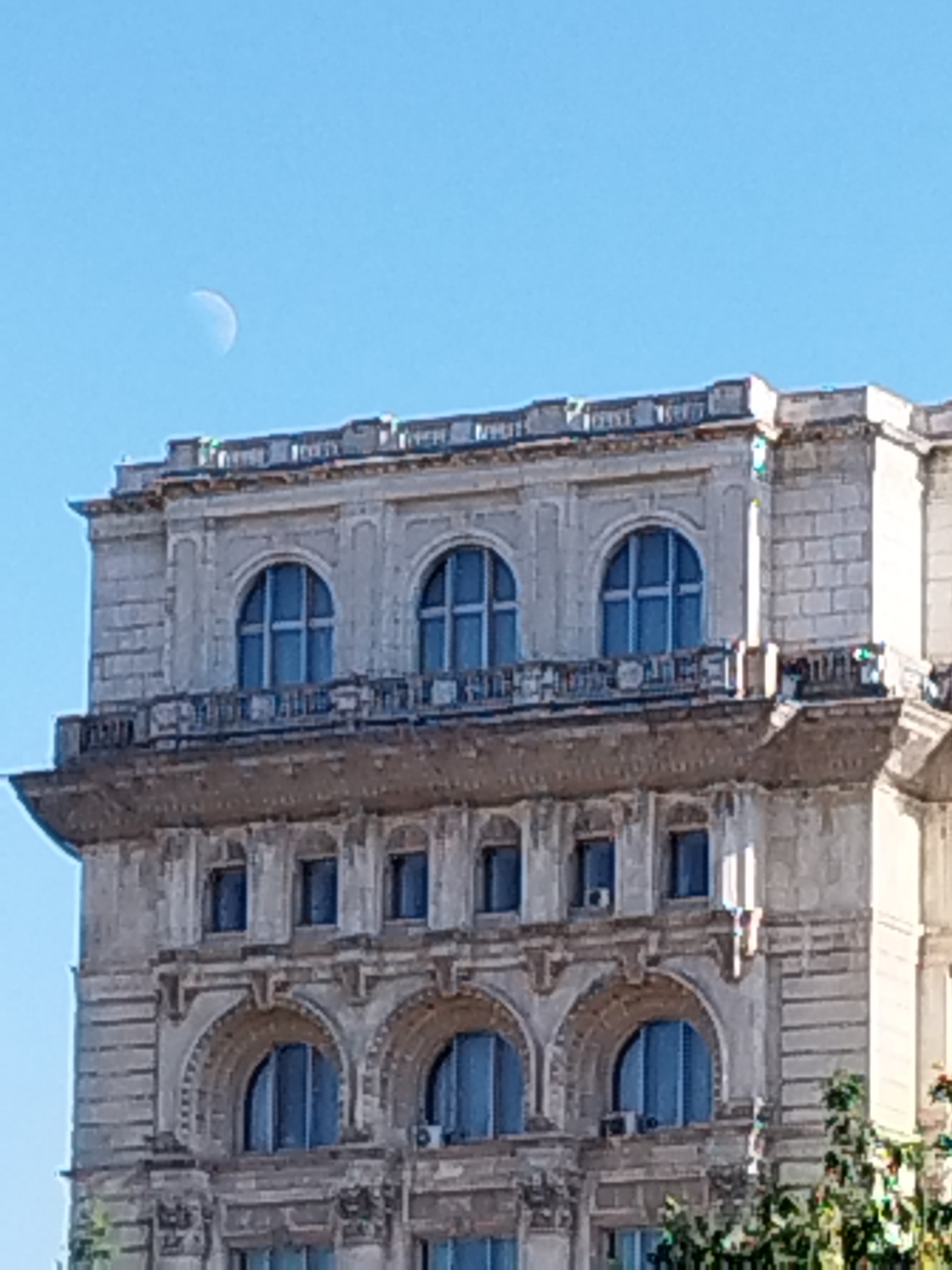 The image shows the upper portion of a historic, ornate building in Bucharest, Romania, against a clear blue sky. The architecture features classical elements, including arched windows with intricate detailing, decorative cornices, and a balustrade. A crescent moon is visible in the sky above the building. The building appears to be made of stone or concrete, with a weathered, aged appearance. The lower part of the frame shows some greenery, suggesting trees or other foliage at the base of the structure. The scene is static and captures a daytime view of the cityscape.