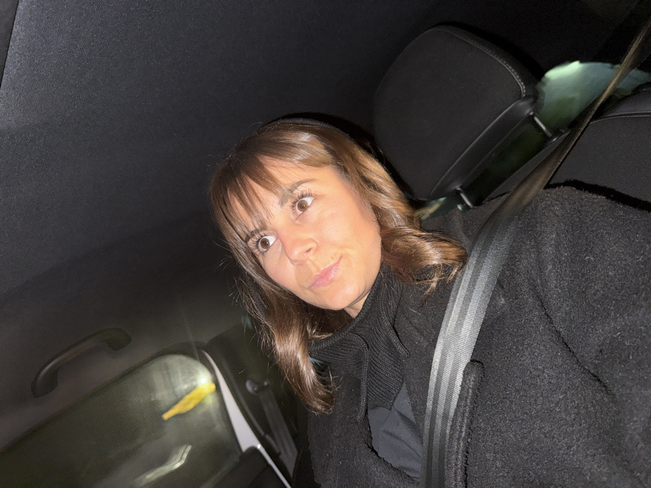A woman with brown hair and bangs, wearing a black coat and scarf, is in the passenger seat of a car. She is wearing a seatbelt. Her eyes are wide and her mouth is slightly pursed, giving a surprised or curious expression. The interior of the car is dark, with a black ceiling and a window visible in the background. The headrest of the driver's seat is visible behind her. The lighting is somewhat dim, emphasizing the woman's face.