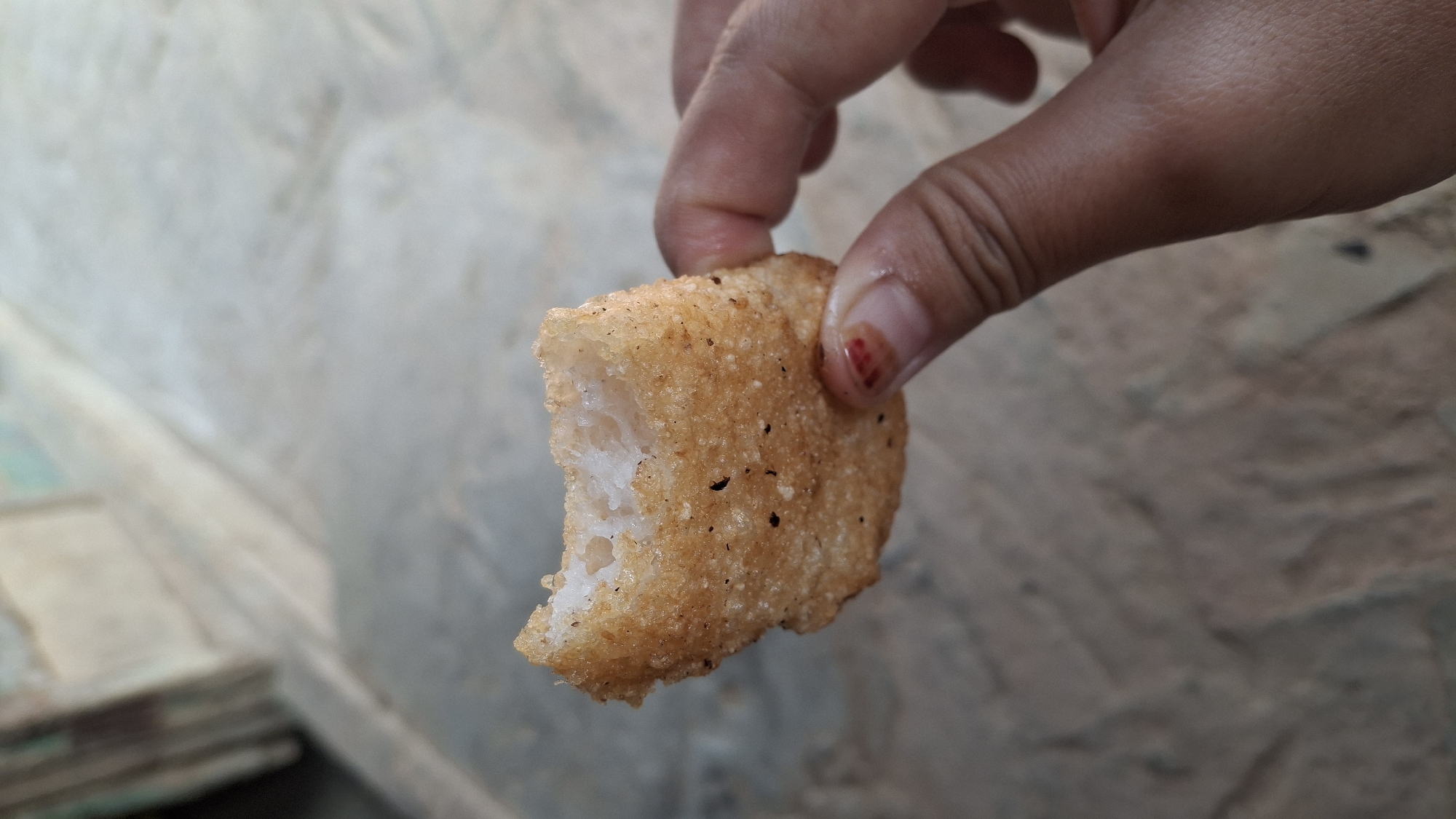 A person's hand holds a partially eaten, fried food item against a blurred background that appears to be an outdoor, possibly urban, setting with some textured ground. The food item is roughly rectangular with a crispy, golden-brown exterior speckled with dark bits, suggesting seasoning or char. The interior is white and appears to have a fluffy or crumbly texture. The hand is holding the food item by the side, with fingers curled around it. The person's thumb is visible, and there's a small reddish mark on the nail of the index finger. The background is out of focus, showing a mix of muted colors and textures, with what could be a wall or building element on the left side. The overall impression is a casual moment of eating outdoors. The location is Pucakwangi, Indonesia.