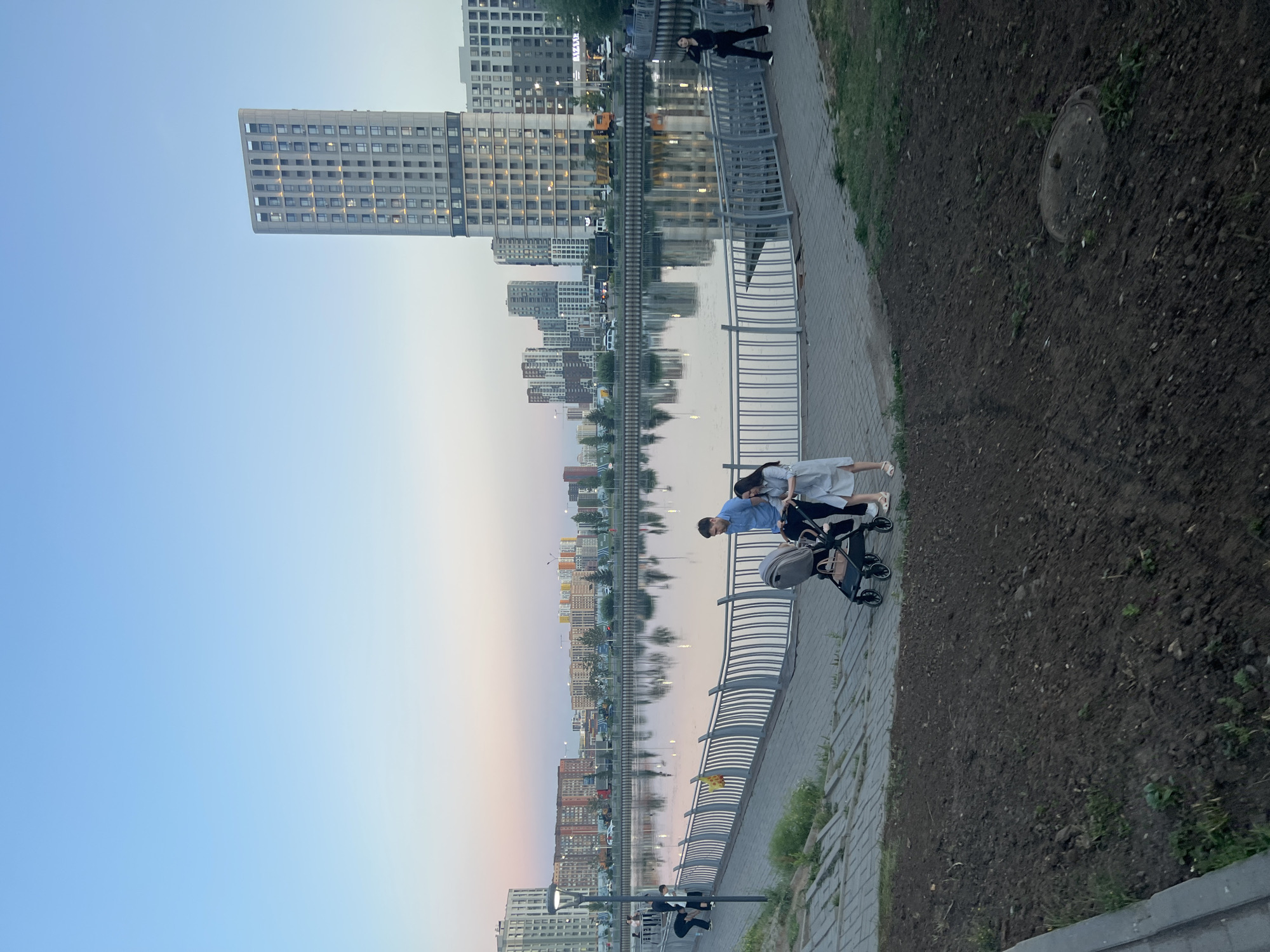 The image depicts a waterfront promenade in Astana, Kazakhstan, with a family pushing a stroller. The scene is set during dusk, with a clear blue sky transitioning into soft pastel hues near the horizon. Modern high-rise buildings, characteristic of Astana's architecture, dominate the background, their reflections shimmering on the calm water. The promenade itself is paved with grey bricks, and a metal railing separates it from the waterfront. A young couple, dressed casually, are the central figures, their attention focused on the stroller. Other individuals are visible further along the promenade and in the distance, suggesting a public gathering space. The foreground shows a patch of dark soil with some sparse greenery, indicating an area of landscaping. The overall atmosphere is serene and urban.