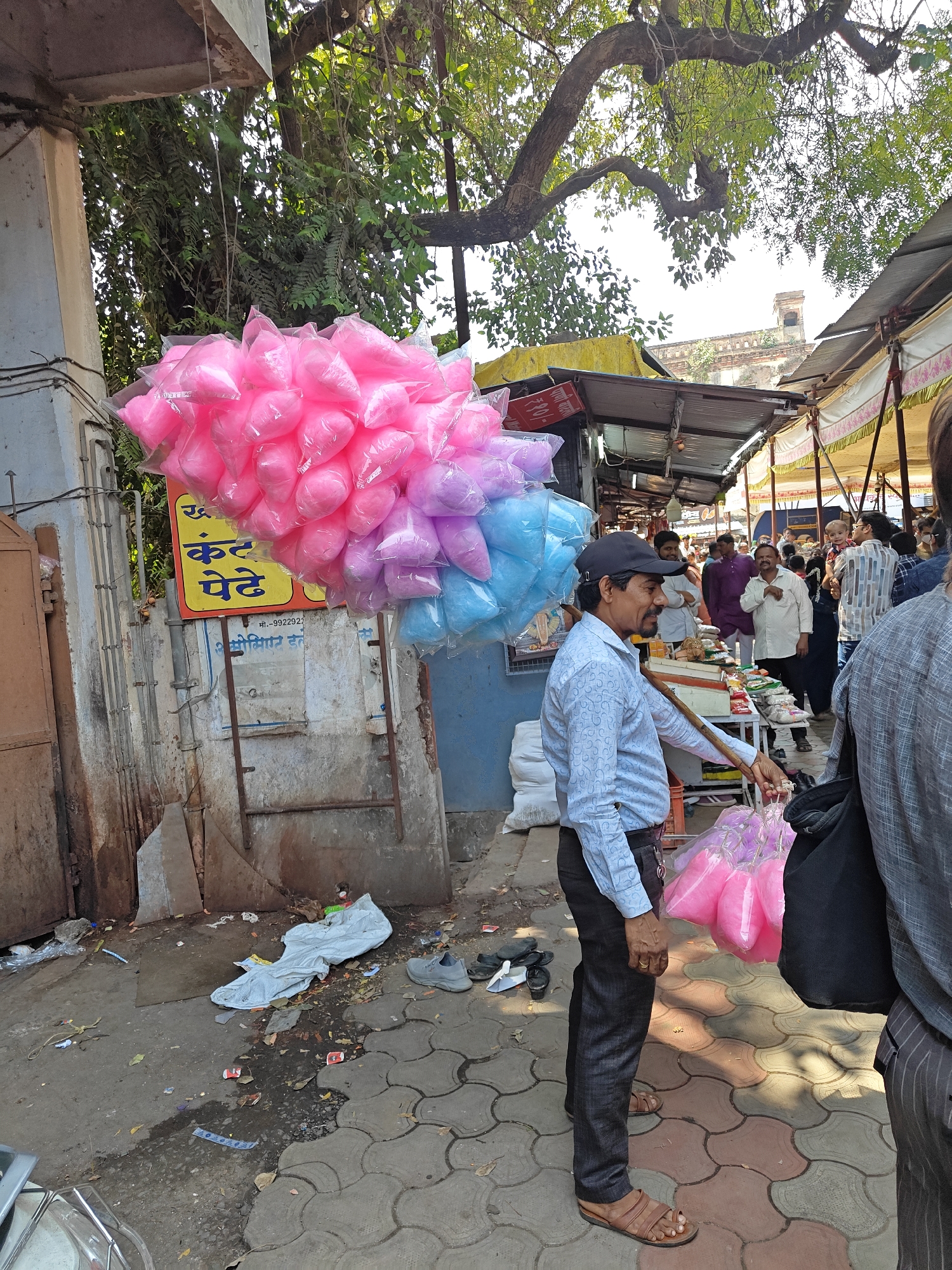 This image depicts an outdoor market scene in the city of Unknown, Unknown, under bright daylight conditions.
In the foreground, a male vendor, possibly in his 40s or 50s, stands facing slightly left, holding a long stick from which numerous clear plastic bags of pink cotton candy are suspended. He wears a light blue long-sleeved shirt with a subtle pattern, dark trousers, brown leather sandals, and a dark baseball cap. A small black pouch is clipped to his belt. Another large cluster of pink, light purple, and light blue cotton candy bags hangs from an unseen structure to his left. The back of another individual, wearing a grey striped shirt and a black backpack, is partially visible in the lower right foreground.
Behind the vendor, a bustling market street extends into the background, lined with numerous makeshift stalls featuring corrugated metal or tarp roofs. Multiple individuals are visible along the street, walking and congregating near the stalls. One person in the background holds a small child.
To the immediate left of the vendor, a grey concrete pillar with electrical conduits attached is visible, alongside a partially open, rusty brown metal door. A yellow sign with text in a local script (identifying "कंठ पेठे" and "₹10/-" for price, and a mobile number "मा.- 9922922") is affixed to a wall or structure behind the vendor. Overhead, a large tree with a thick, gnarled trunk and green foliage extends across the upper portion of the frame. In the far background, an older brick building with a distinct tower or spire is visible above the market roofs.
The ground consists of a mixture of grey and reddish interlocking paving stones and bare dirt, strewn with various litter including plastic bags, wrappers, and discarded footwear (a pair of grey shoes and black sandals). Shadows on the ground suggest the sun is high.