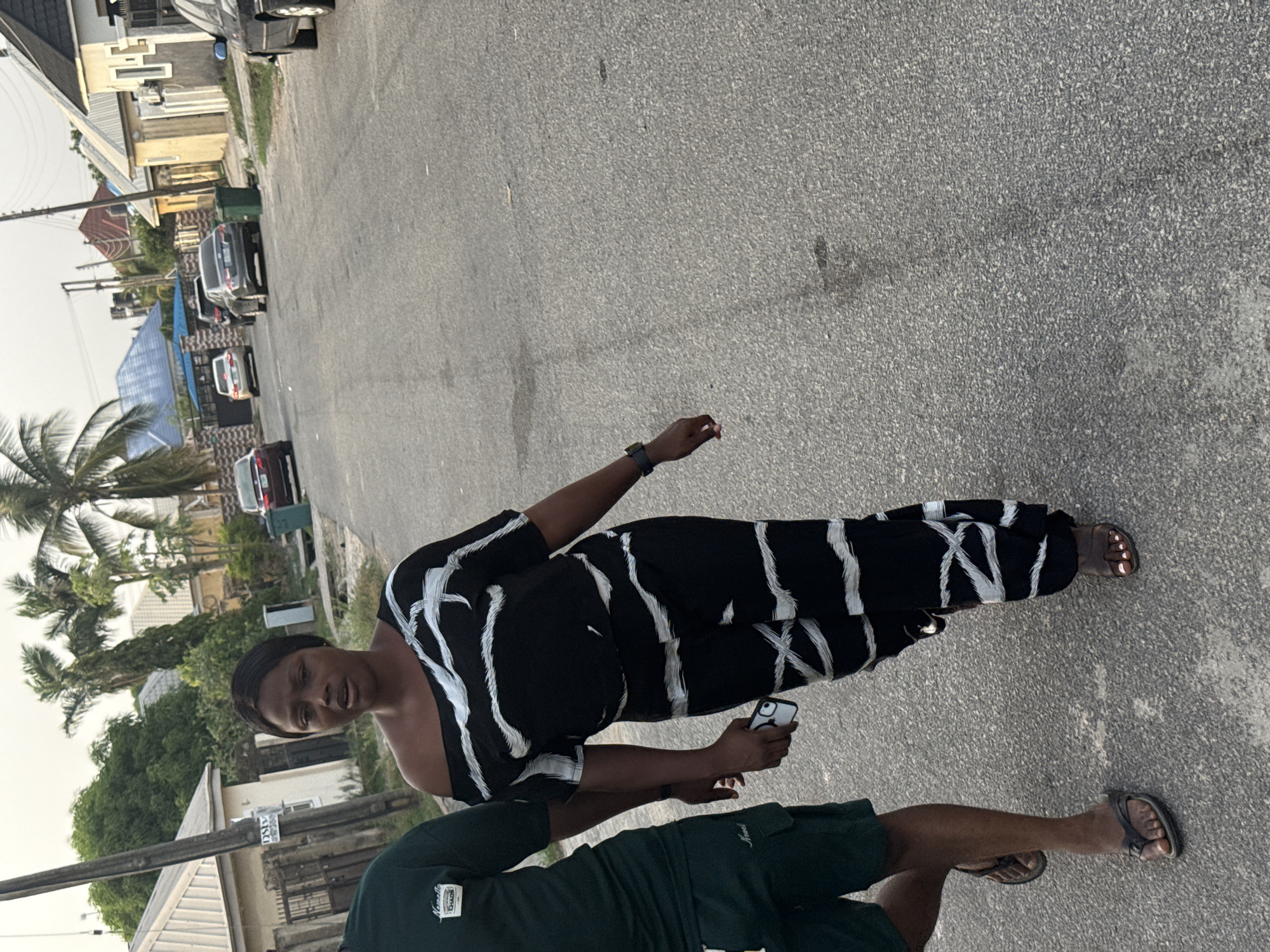 The image captures a street scene in a residential area, consistent with Abuja, Nigeria, during daylight hours. The composition is rotated, showing two individuals on a paved road.
In the mid-ground, a woman is prominent, facing generally forward. She has dark hair and wears a black off-shoulder top paired with wide-leg pants, both featuring white abstract vertical and diagonal stripe patterns. A dark smartwatch is on her left wrist, and she holds a white smartphone in her right hand. She wears light-colored open-toed sandals. Her expression is neutral, and she appears to be looking slightly off-camera.
Partially visible in the immediate foreground, largely obscuring the woman's lower half, is another person. They wear a dark green short-sleeved top with a white "Arcade" logo and other indistinct text on the chest, along with dark green shorts and dark-colored sandals.
The background reveals a paved asphalt street flanked by various houses and buildings. The architecture varies, with some houses featuring light-colored walls and others distinct blue or red roofs. Several cars are parked along the street, including a black SUV, a white sedan, and a red car. Abundant green foliage, including tall palm trees, indicates a lush environment. A green trash bin is visible near the parked vehicles. Utility poles with overhead wires run alongside the street. The sky is bright, suggesting daytime, possibly under an overcast or hazy condition. A sign attached to a utility pole shows partially legible text, including "DSD".