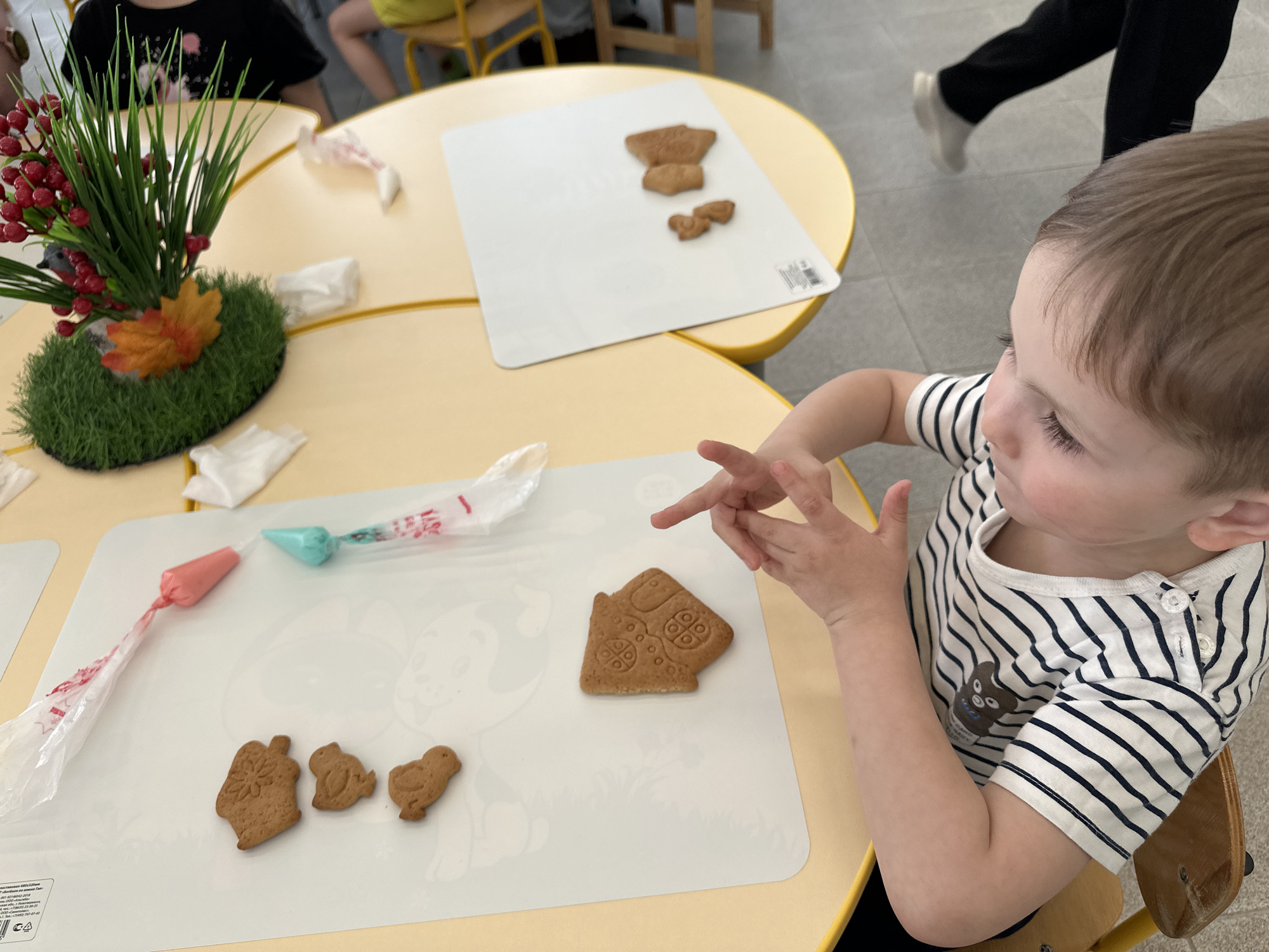 A child is seated at a yellow table decorating gingerbread cookies. The child is wearing a white and navy striped shirt. On the table, there is a white mat with a drawing of a cartoon dog, and several gingerbread cookies shaped like a house, a pumpkin, and two birds. There are two piping bags filled with colored icing on the mat. On the same table, there is a decorative centerpiece of greenery, red berries, and an orange flower. Another table is visible in the background with similar gingerbread cookies on a white mat. A person in black pants and white shoes is walking behind the table.