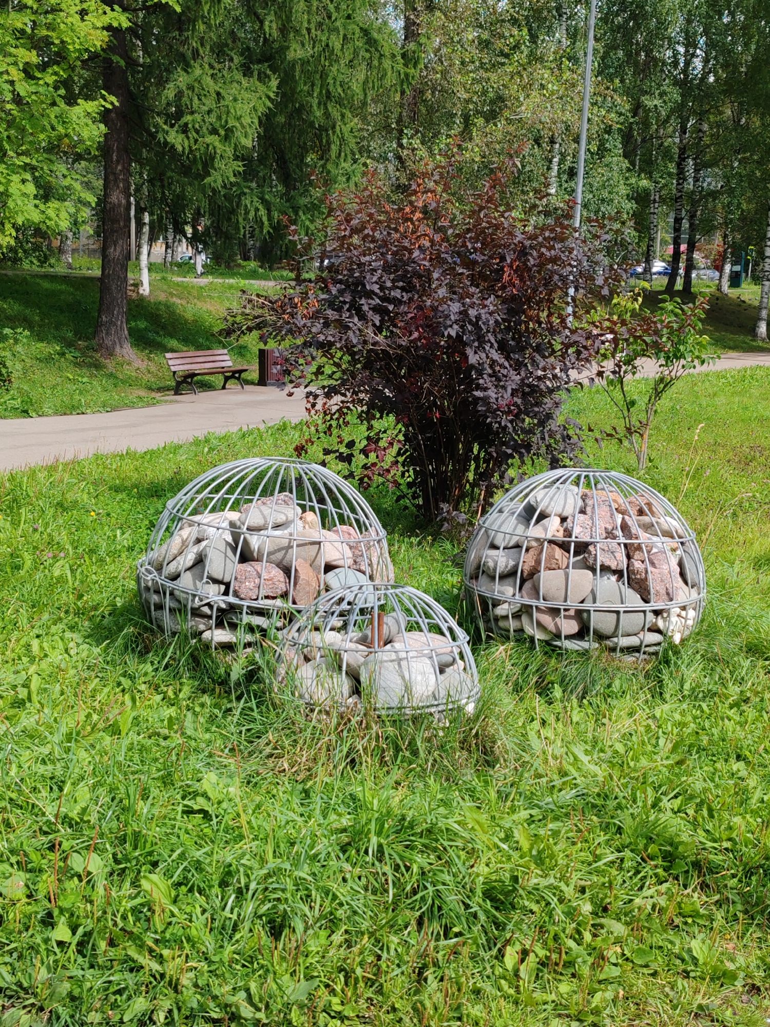 Three spherical metal cages filled with stones are placed on a grassy area in a park. The cages are of varying sizes, with the largest on the right, a medium-sized one in the center, and the smallest in the foreground. Behind the cages, a dark red-leaved bush is prominent. Further in the background, a paved path runs along the edge of the grass, and a wooden bench is situated next to it. Trees with green foliage, some appearing to be birch trees with white bark, form a backdrop. The scene is bathed in sunlight, casting shadows on the grass. The location is identified as Syktyvkar, Russia.
