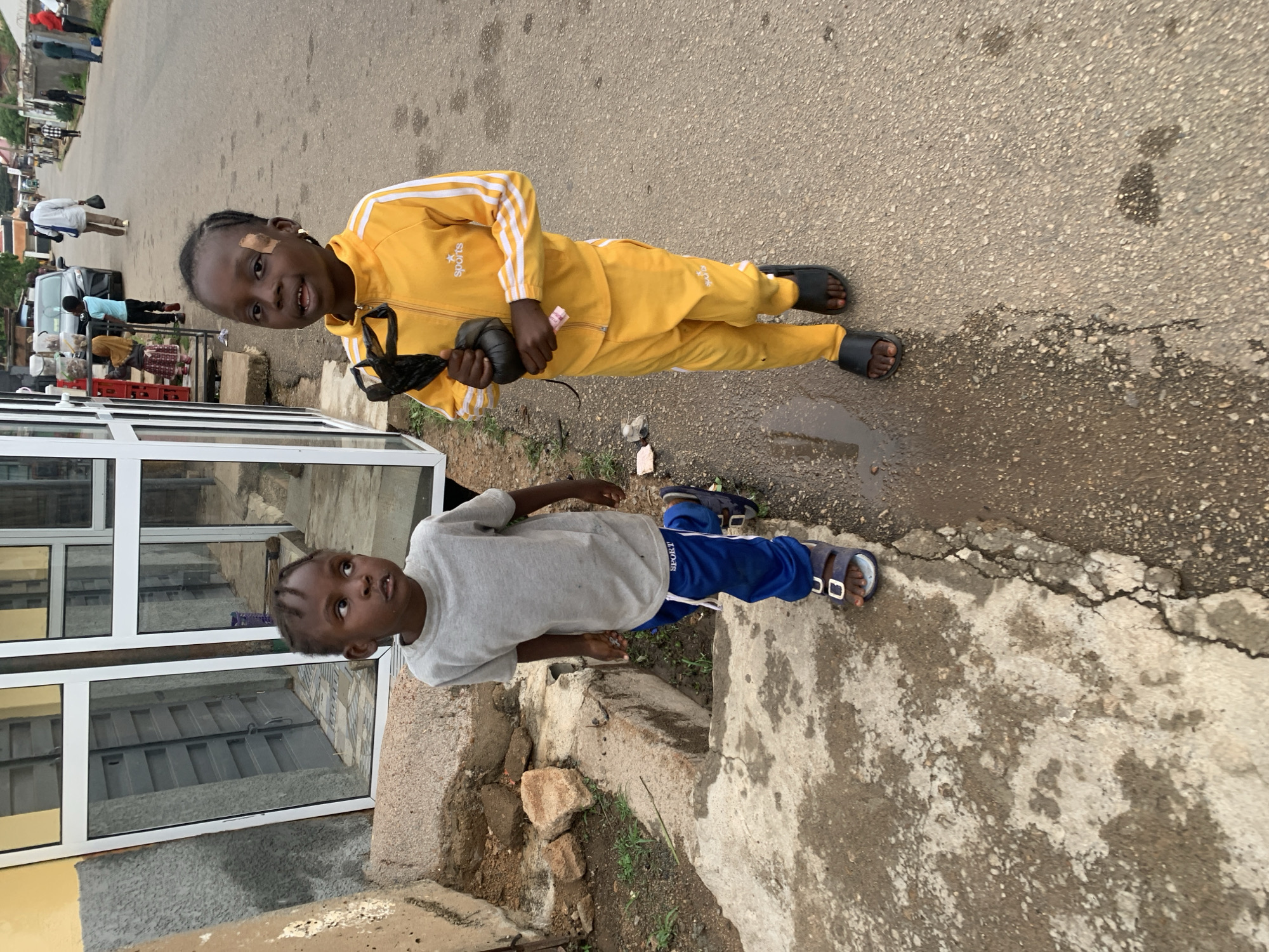 Two young children are standing outside a building in Jos, Nigeria. One child is wearing a yellow tracksuit with white stripes and black sandals, holding a black object. The other child wears a gray t-shirt and blue tracksuit bottoms with sandals. They are standing on or beside a concrete sidewalk. The location seems to be near a shop or a building with large windows. The street and sidewalk show signs of wear and repair. Other people and vehicles are visible in the background.