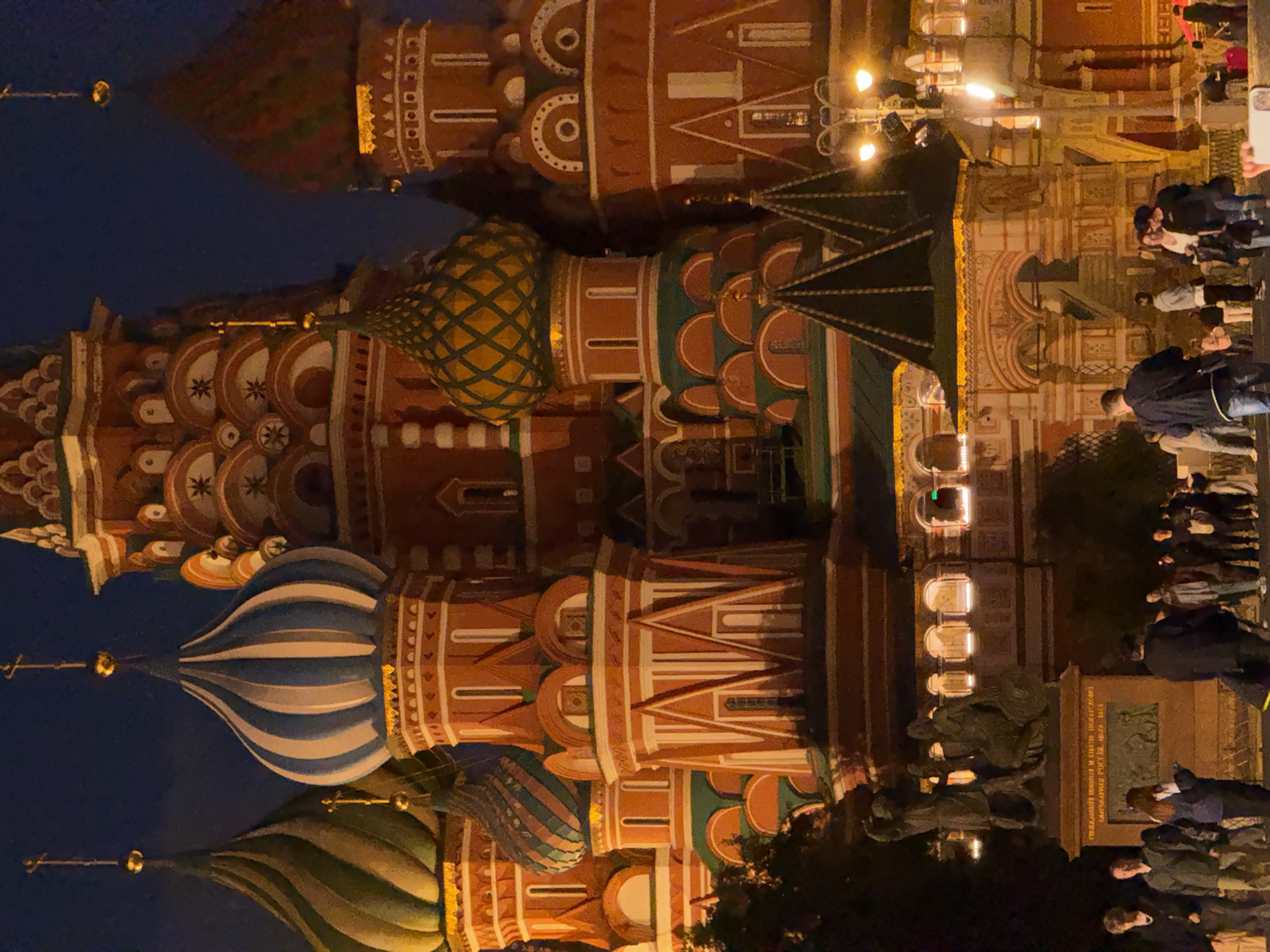 The image depicts Saint Basil's Cathedral in Moscow, Russia, at night. The iconic onion domes of the cathedral, in various colors and patterns, are illuminated by artificial light against a dark blue sky. In the foreground, a crowd of people is gathered on a paved area. Some individuals are standing, others are walking, and a few are engaged in conversation. A statue on a pedestal is visible to the left, and streetlights illuminate the scene. The architecture is ornate and traditional, characterized by red brick walls and colorful embellishments on the domes and towers.